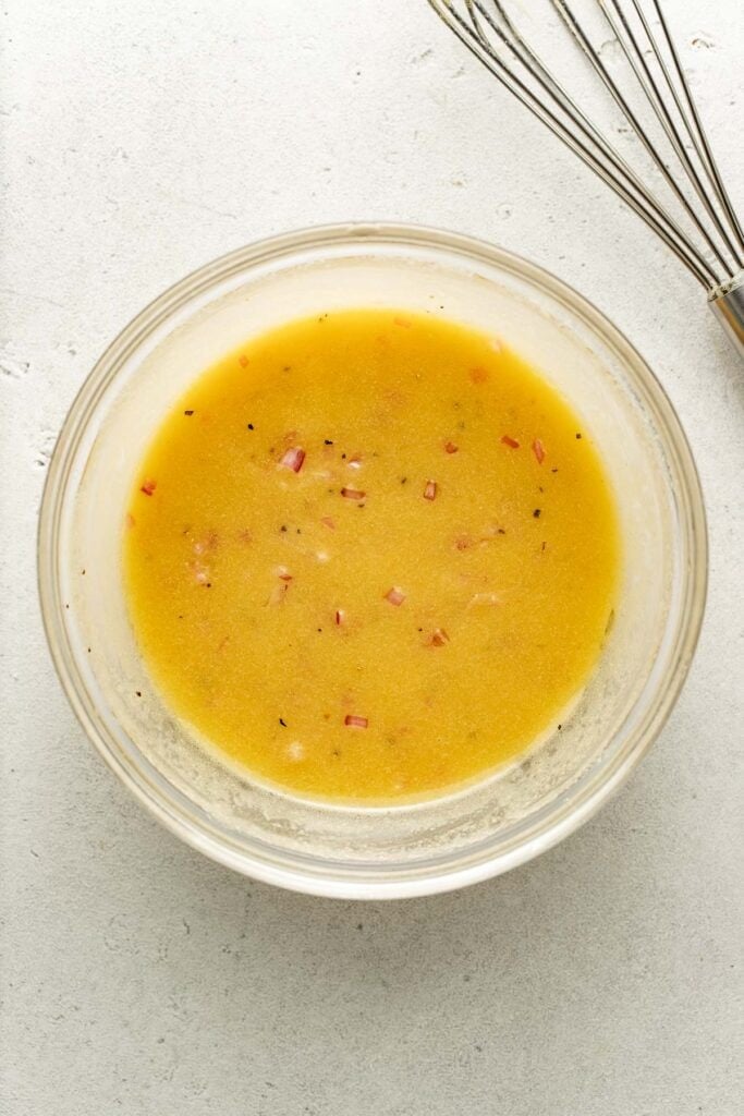 A glass bowl filled with yellow vinaigrette dressing with small pieces of red onion, next to a metal whisk on a light countertop.