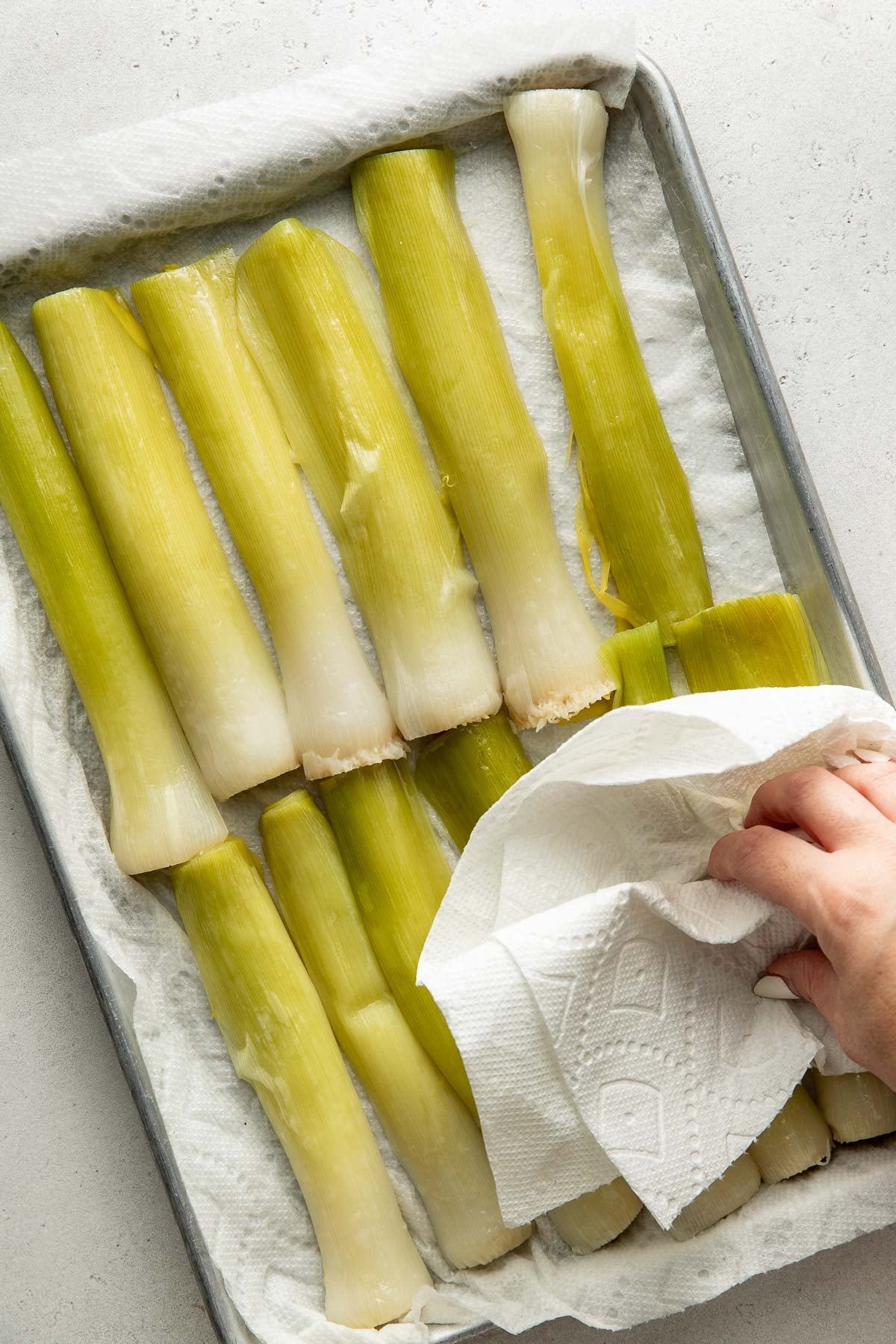 A hand uses a paper towel to dry several cooked leek stalks arranged on a paper towel-lined baking sheet.