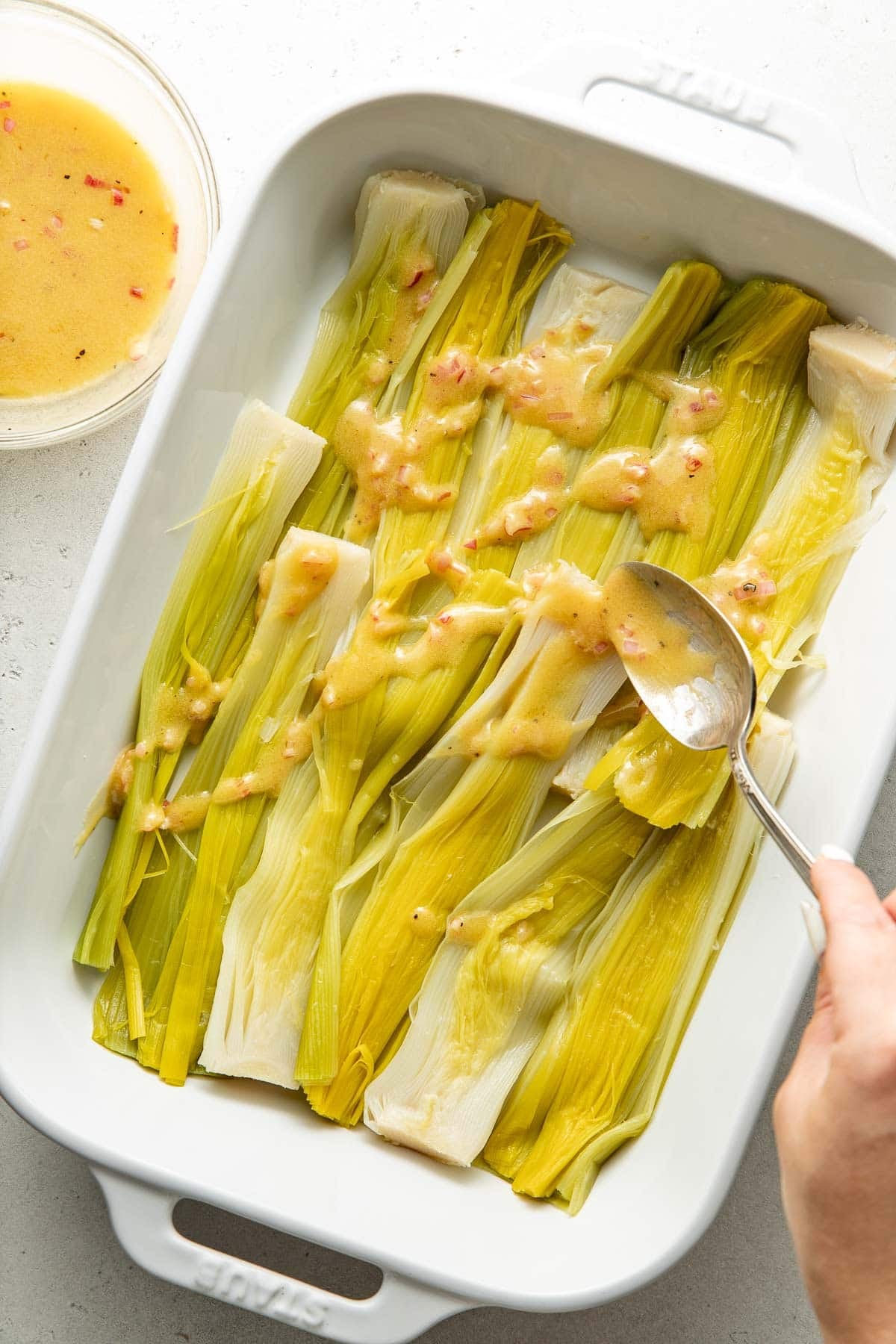 A hand drizzles sauce over cooked leeks arranged in a white baking dish. A bowl with extra sauce sits nearby on a light surface.