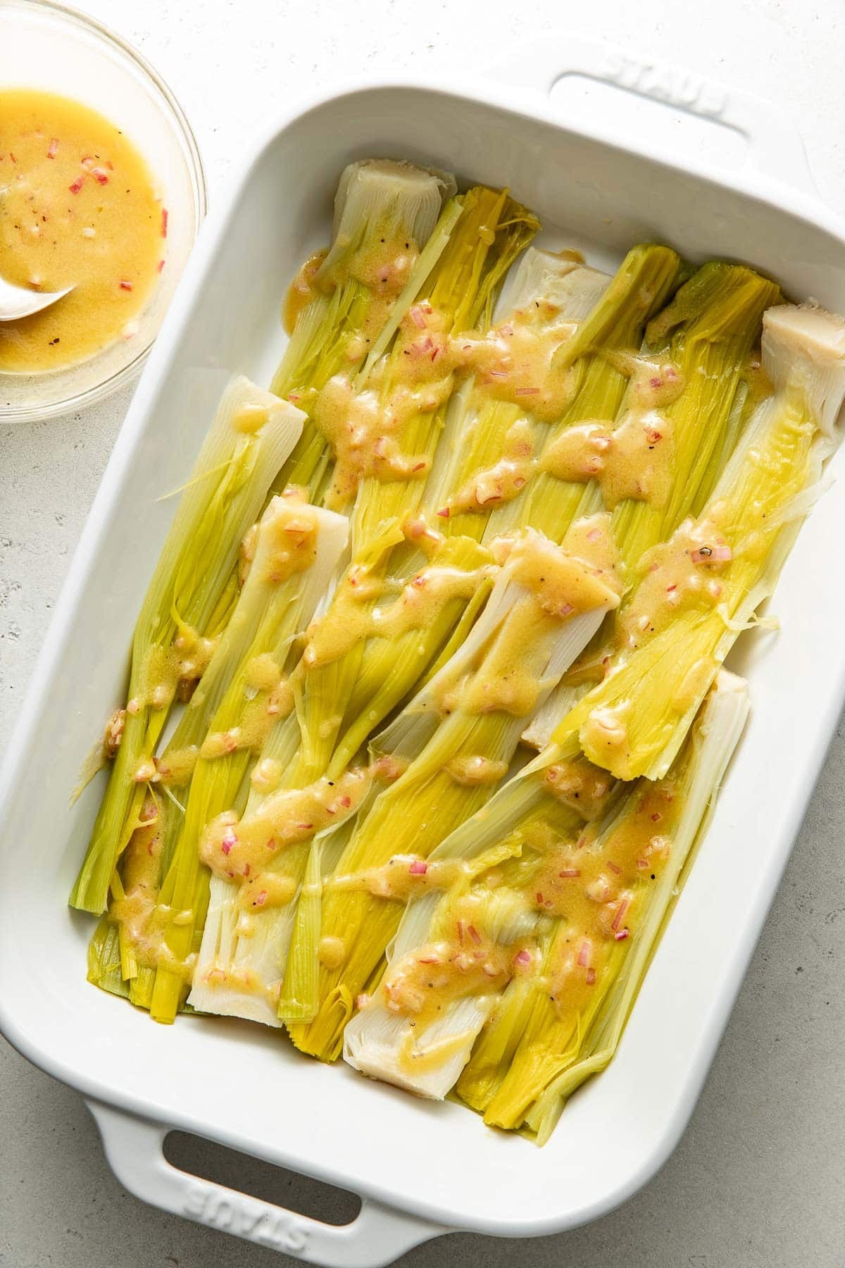 A white baking dish filled with cooked leeks, topped with a vinaigrette dressing containing visible bits of red pepper. A small glass bowl with extra dressing sits nearby on a light surface.