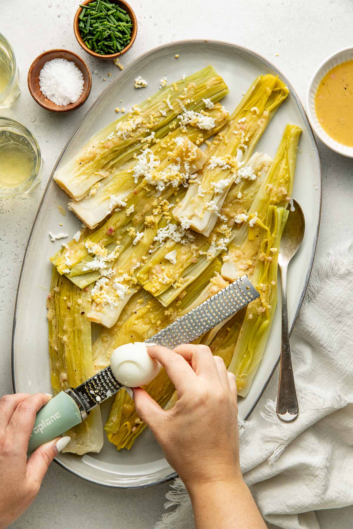 A person grates a hard-boiled egg over cooked leeks on a platter, topped with crumbled cheese, with salt, chives, and a bowl of sauce nearby.