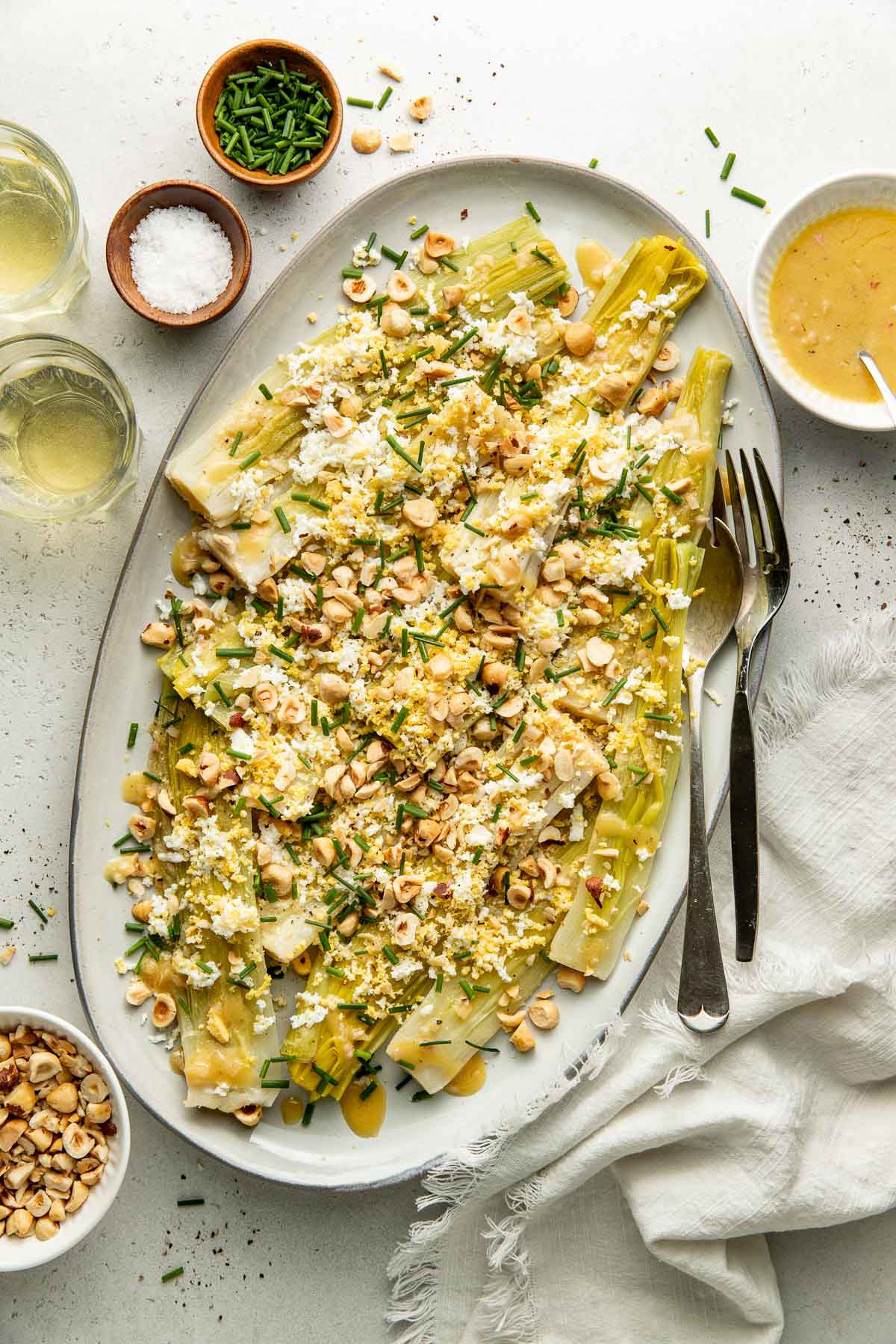 Oval platter of leeks topped with crumbled cheese, chopped hazelnuts, and herbs. Two glasses of white wine, small bowls of salt, nuts, herbs, and dressing are nearby, with serving utensils and a napkin beside the platter.