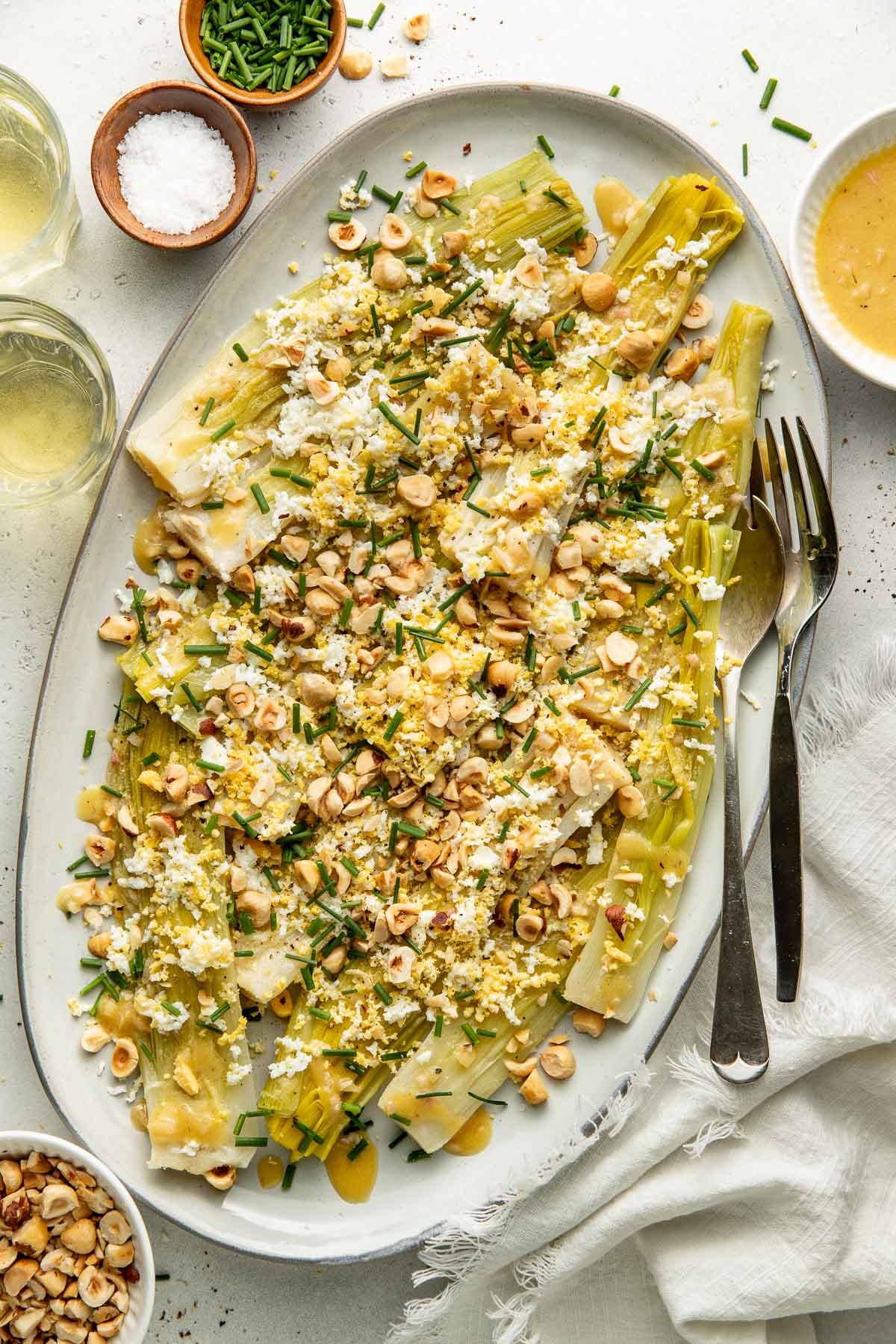 Oval platter filled with cooked leeks topped with chopped hazelnuts, crumbled cheese, grated egg, and chopped chives. Two forks, a knife, bowls of salt, nuts, sauce, and glasses of white wine are nearby.