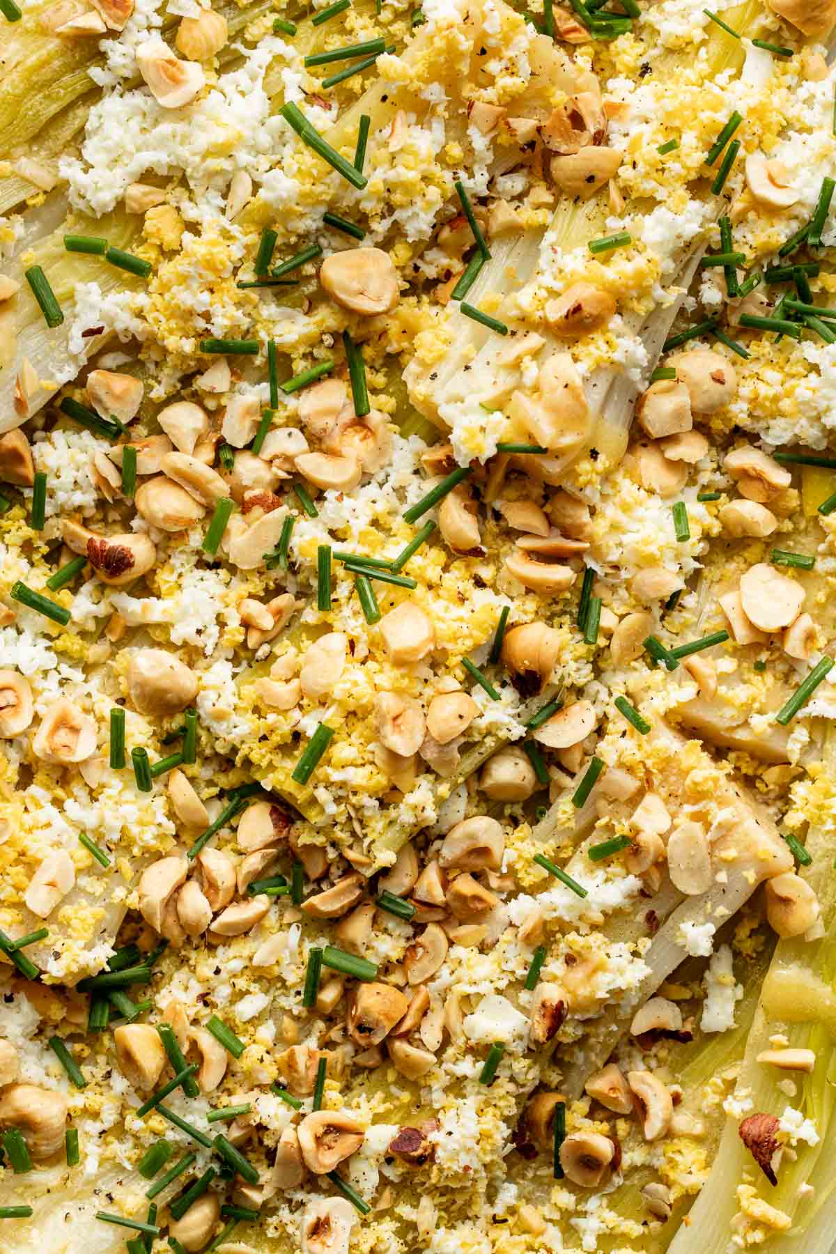 Close-up of a dish topped with chopped hazelnuts, grated cheese or egg, and chopped chives over vegetables. The texture is crunchy and creamy with green, yellow, and beige tones.
