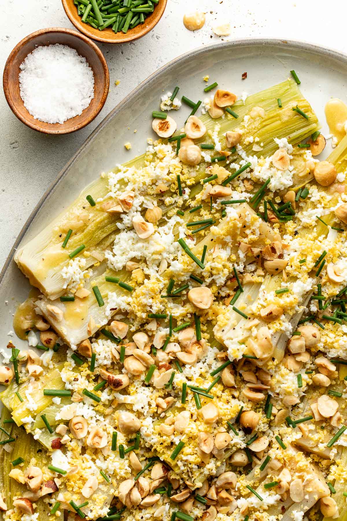 Leeks topped with chopped hard-boiled eggs, toasted hazelnuts, and fresh chives are arranged on a white platter. Nearby are small bowls of coarse salt and chopped chives.