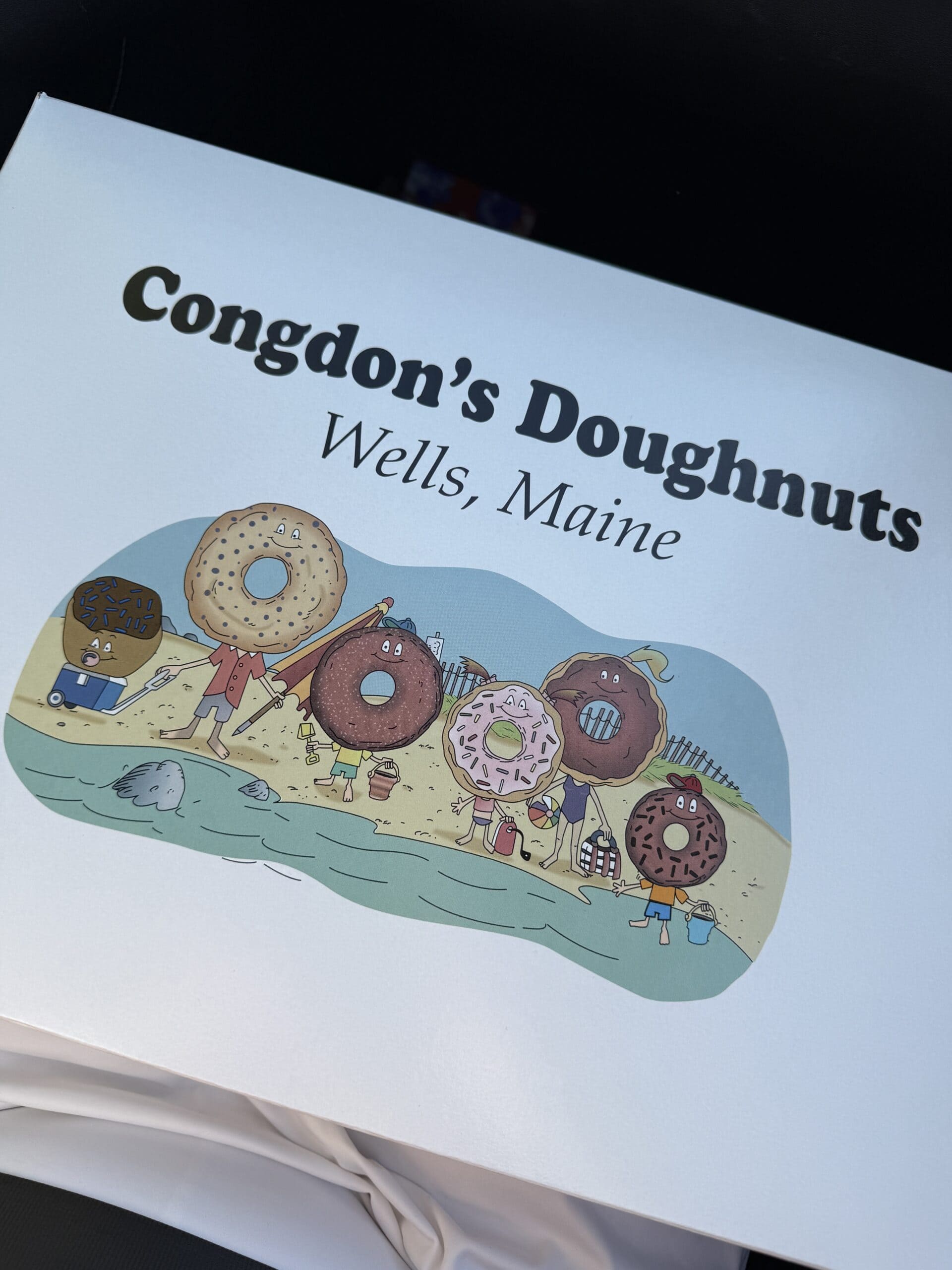 A box with Congdons Doughnuts, Wells, Maine written on it, featuring cartoon doughnuts with faces and arms standing on a beach.