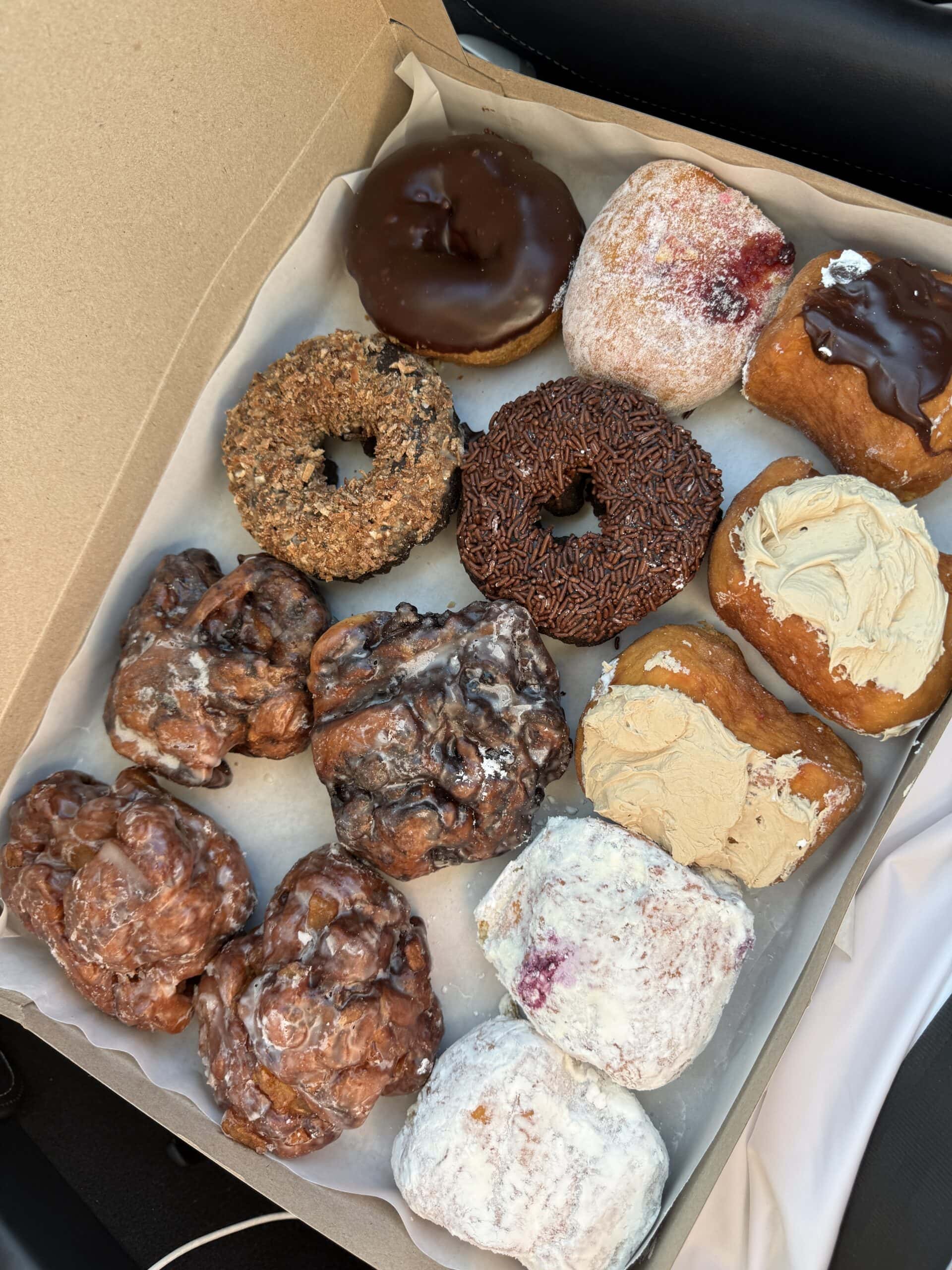 A box of assorted donuts and pastries, including chocolate frosted donuts, sprinkle donuts, cake donuts, filled donuts, apple fritters, and powdered jelly donuts, arranged neatly on parchment paper.