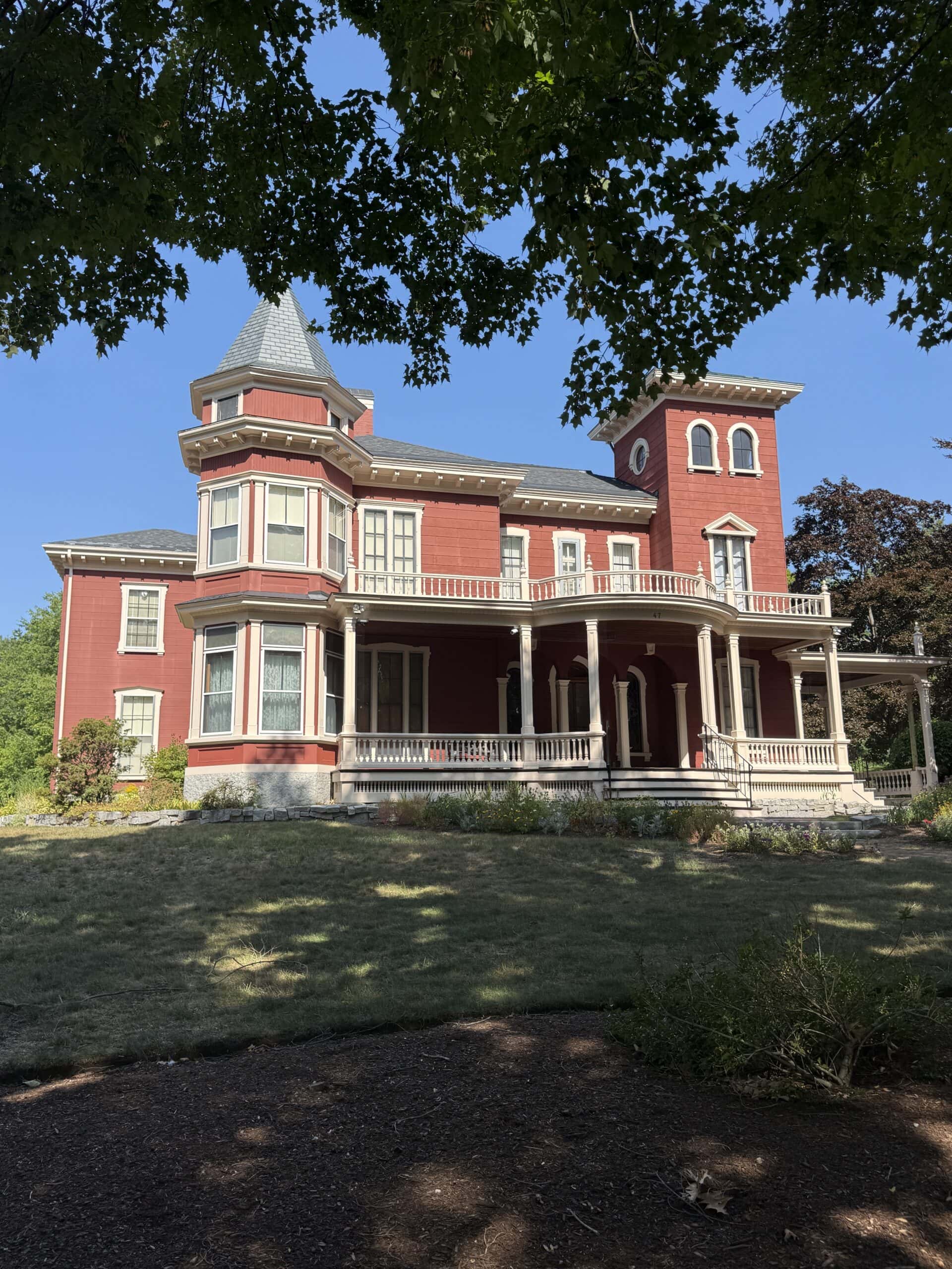 A large red Victorian-style house with a wraparound porch, turret, and tower, set on a grassy yard with trees providing shade in the foreground.