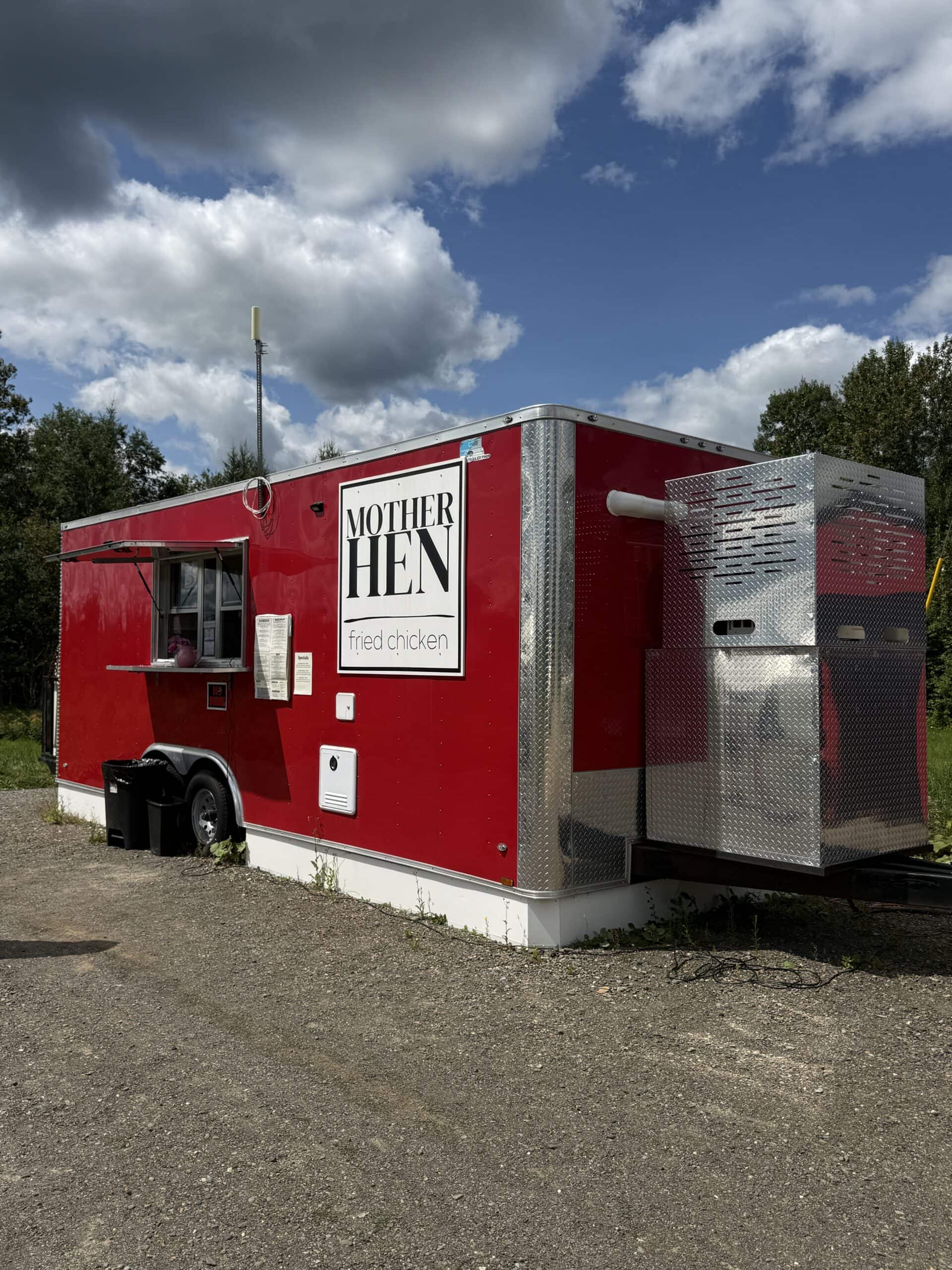 A red food truck labeled Mother Hen Fried Chicken sits on gravel, with trash bins beside it. Trees and a cloudy sky are in the background.