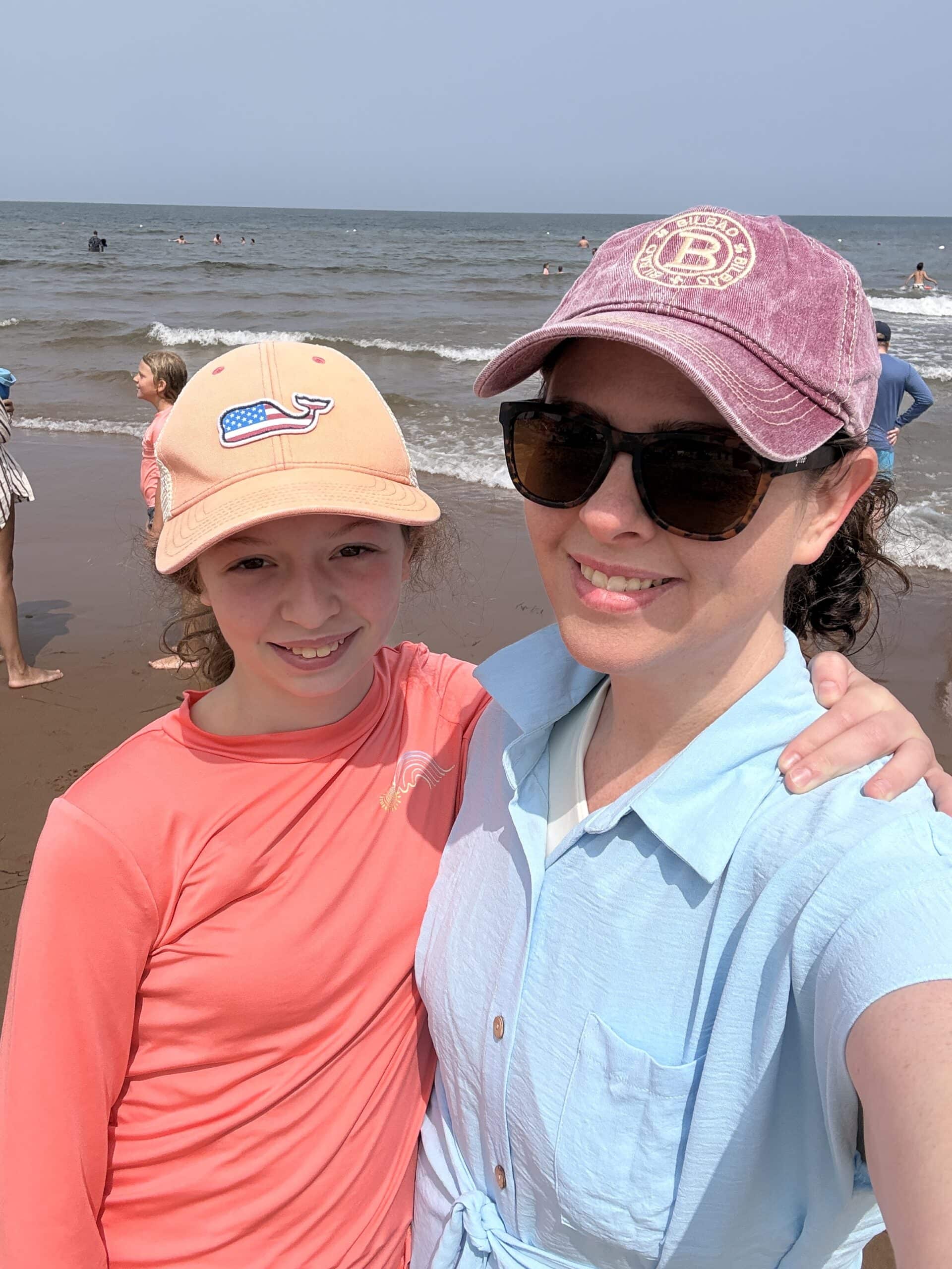 A woman and a girl smile for a selfie on a sunny beach. Both wear hats and sunglasses, with the girl in a peach shirt and the woman in a blue top. Waves and people are visible in the background.