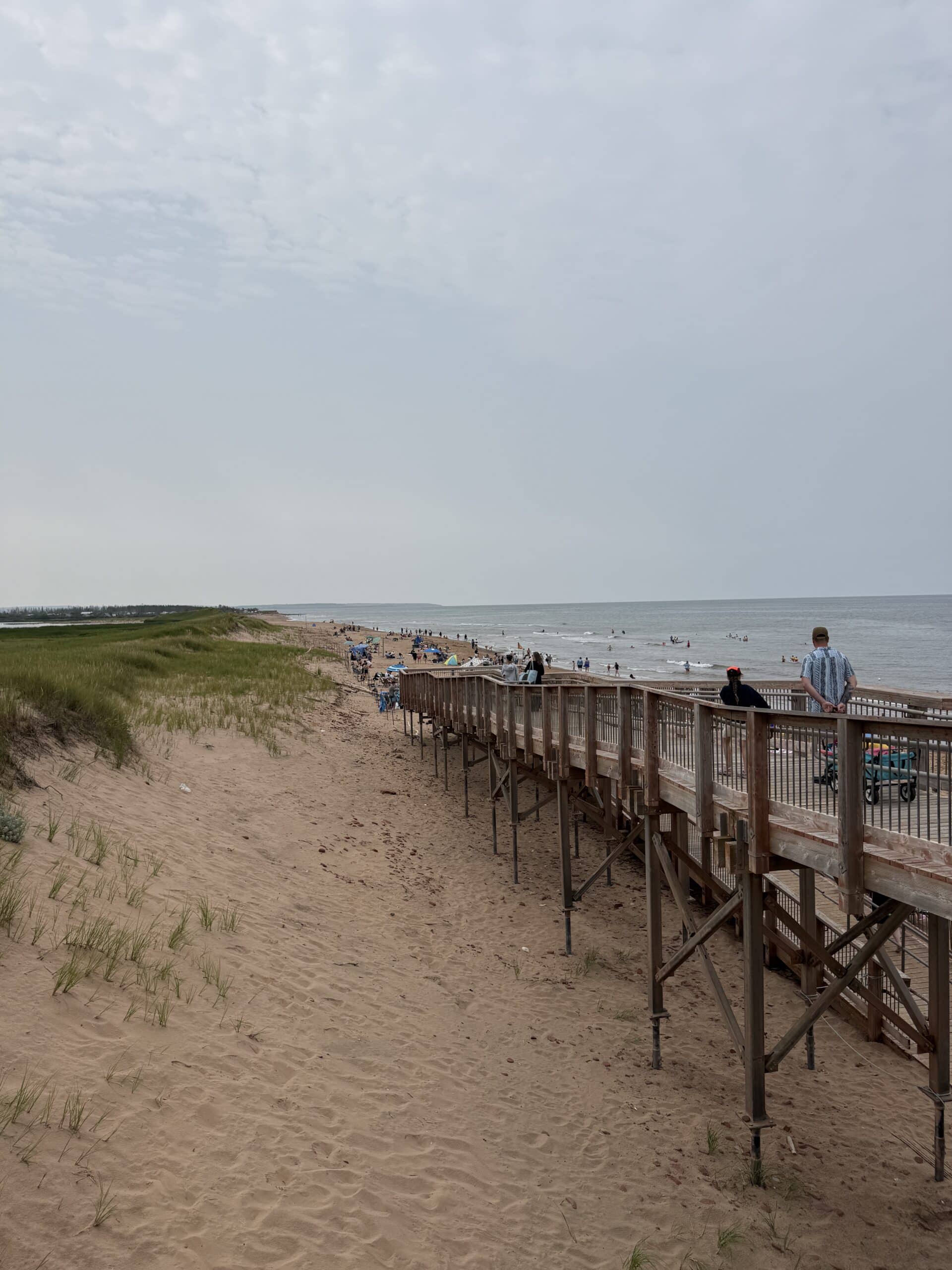 A sandy beach with tall grass, a wooden boardwalk, and people walking or relaxing along the shore under a cloudy sky. The ocean is calm, and more people are visible in the distance.