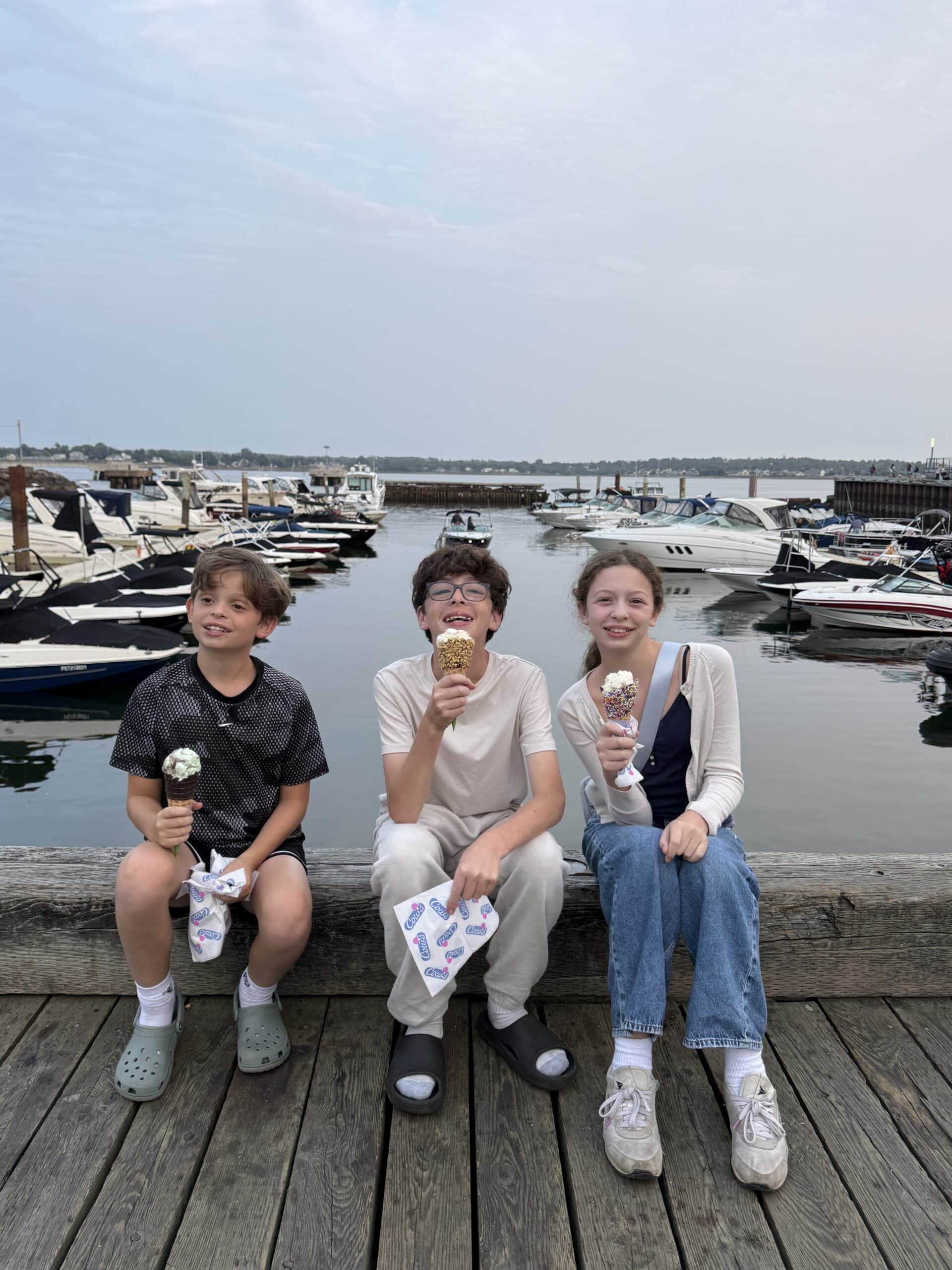 Three kids sit on a wooden dock eating ice cream cones, smiling, with boats and water in the background under a cloudy sky.