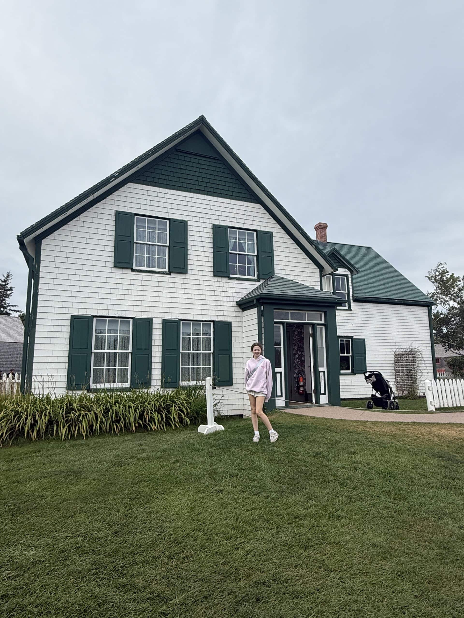 A person in a pink hoodie stands on the lawn in front of a large white house with green trim and a green roof. A stroller is near the open front door. The sky is overcast.