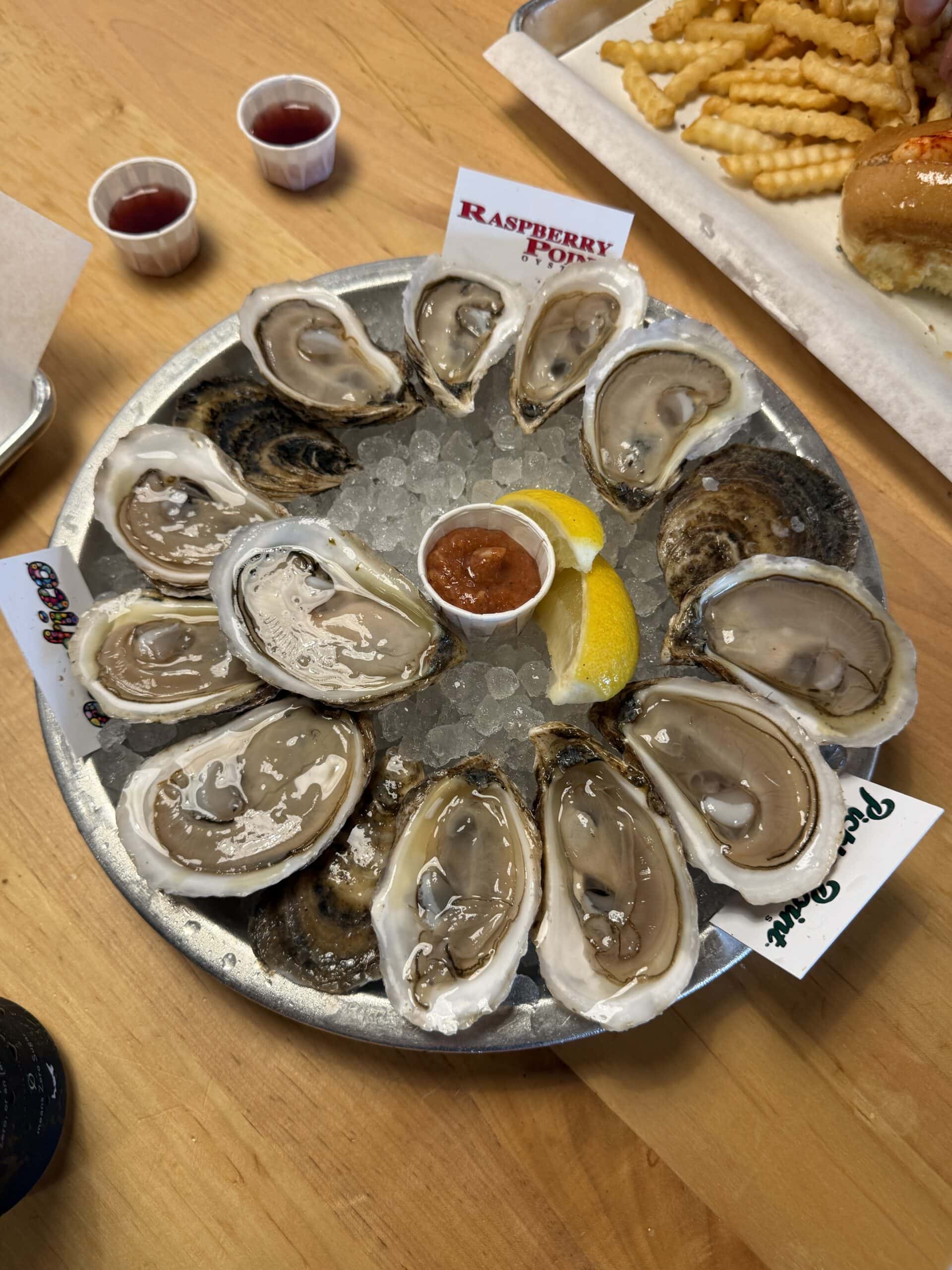 A metal tray of raw oysters on ice with lemon wedges and cocktail sauce in the center. Two labeled oyster varieties are visible. Crinkle-cut fries and a sandwich are in the background.