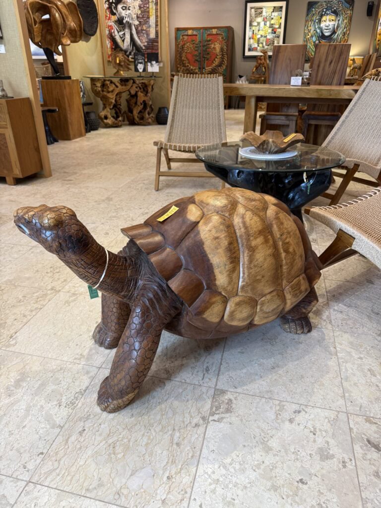 A large, realistic wooden tortoise sculpture sits on a tiled floor in a furniture store, surrounded by chairs, tables, and colorful artwork on the walls.