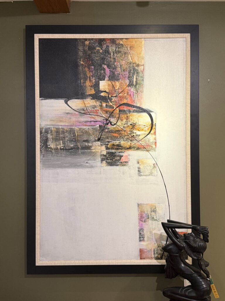 Abstract painting with black, gold, pink, and white colors, featuring bold brushstrokes and a looping black line. A dark metallic sculpture of a person sitting with arms raised is partially visible in the lower right corner.