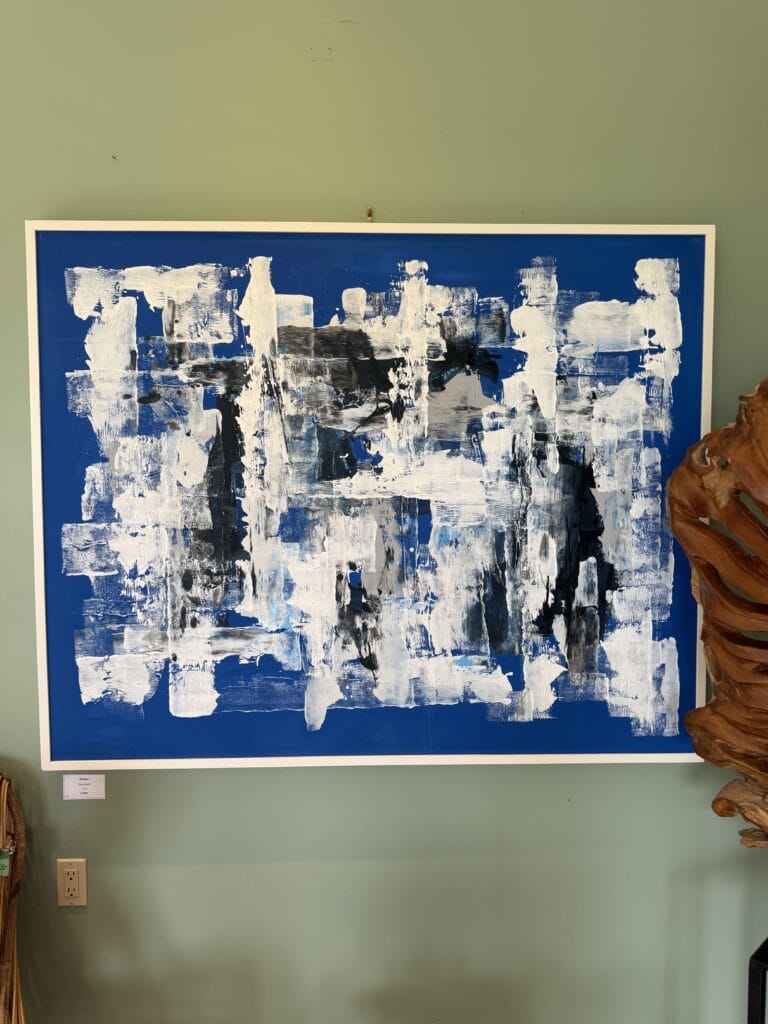 Abstract painting with bold white, black, and blue brushstrokes on a solid blue background, framed in white, hanging on a green wall with a wooden sculpture partially visible on the right.