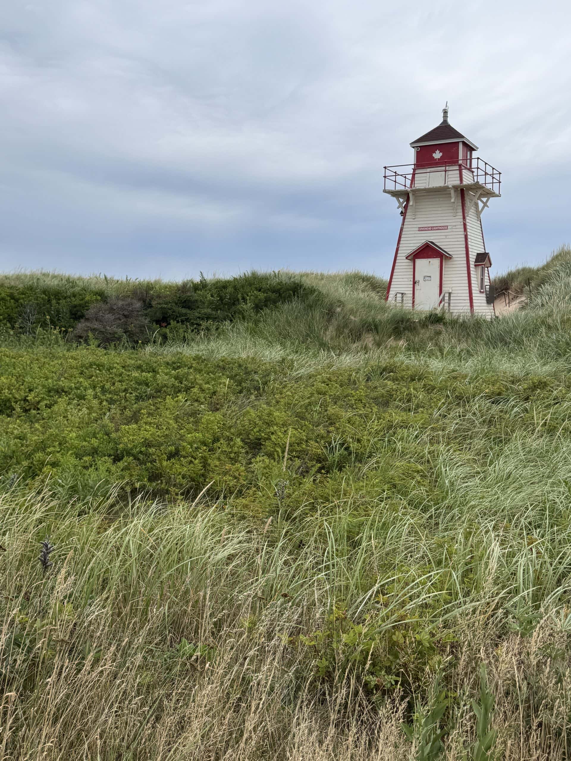 A small white lighthouse with red trim stands amid tall green grass and sand dunes under a cloudy sky.