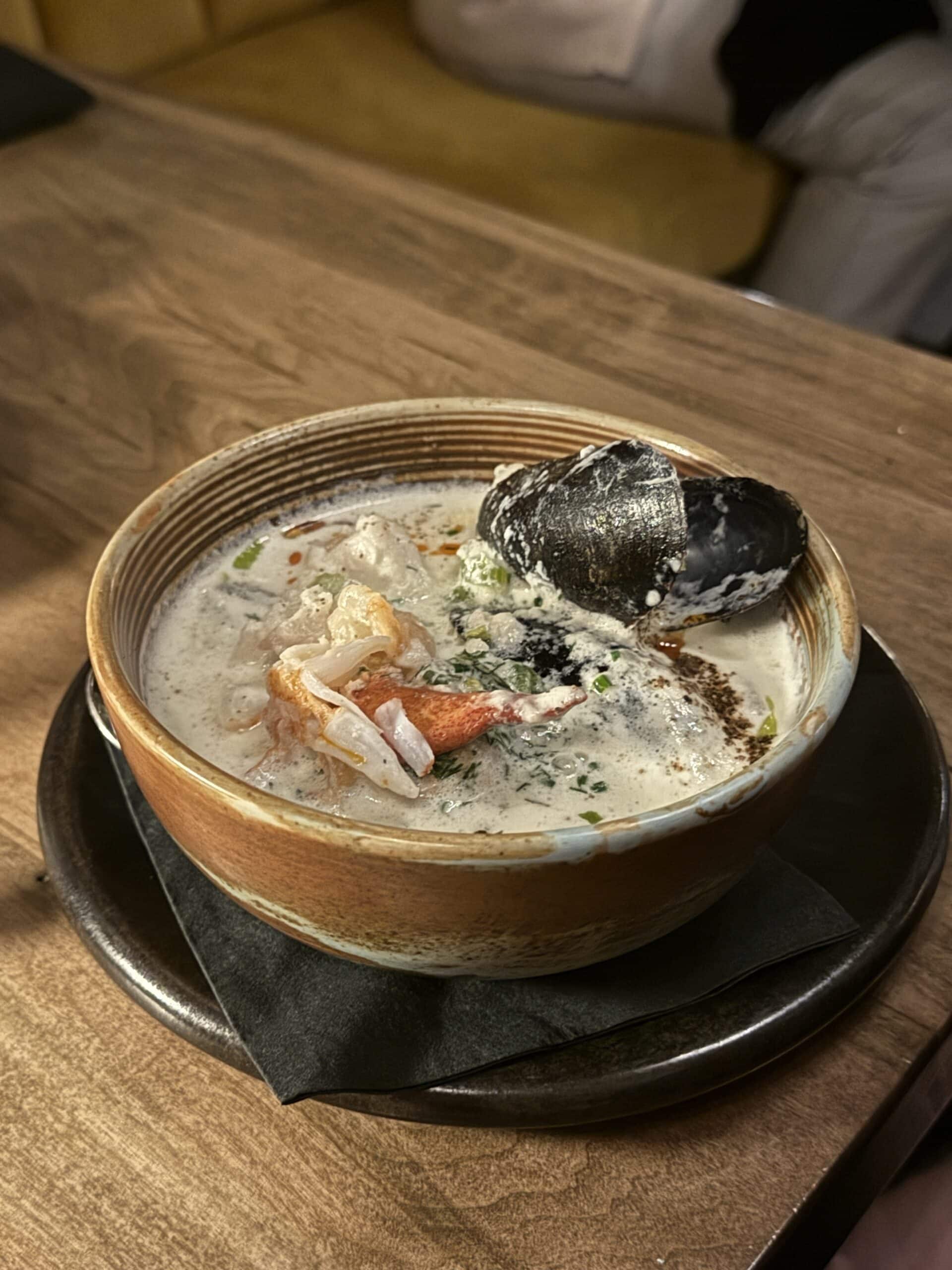 A brown bowl of creamy seafood soup containing a mussel, shrimp, and herbs sits on a black plate atop a wooden table.