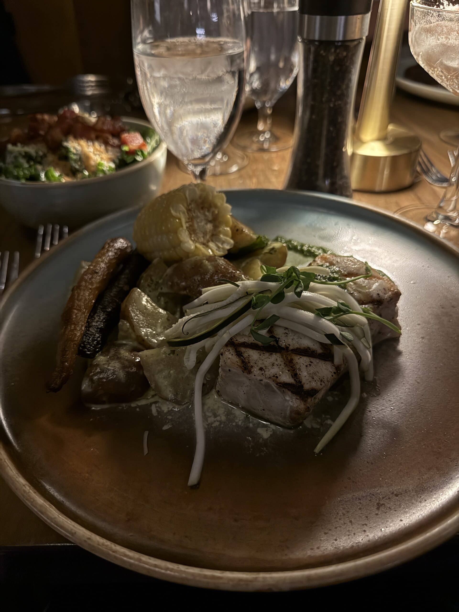 Grilled fish topped with bean sprouts, served with roasted potatoes, corn on the cob, and a roasted carrot on a plate. In the background are glasses of water and a bowl of salad.