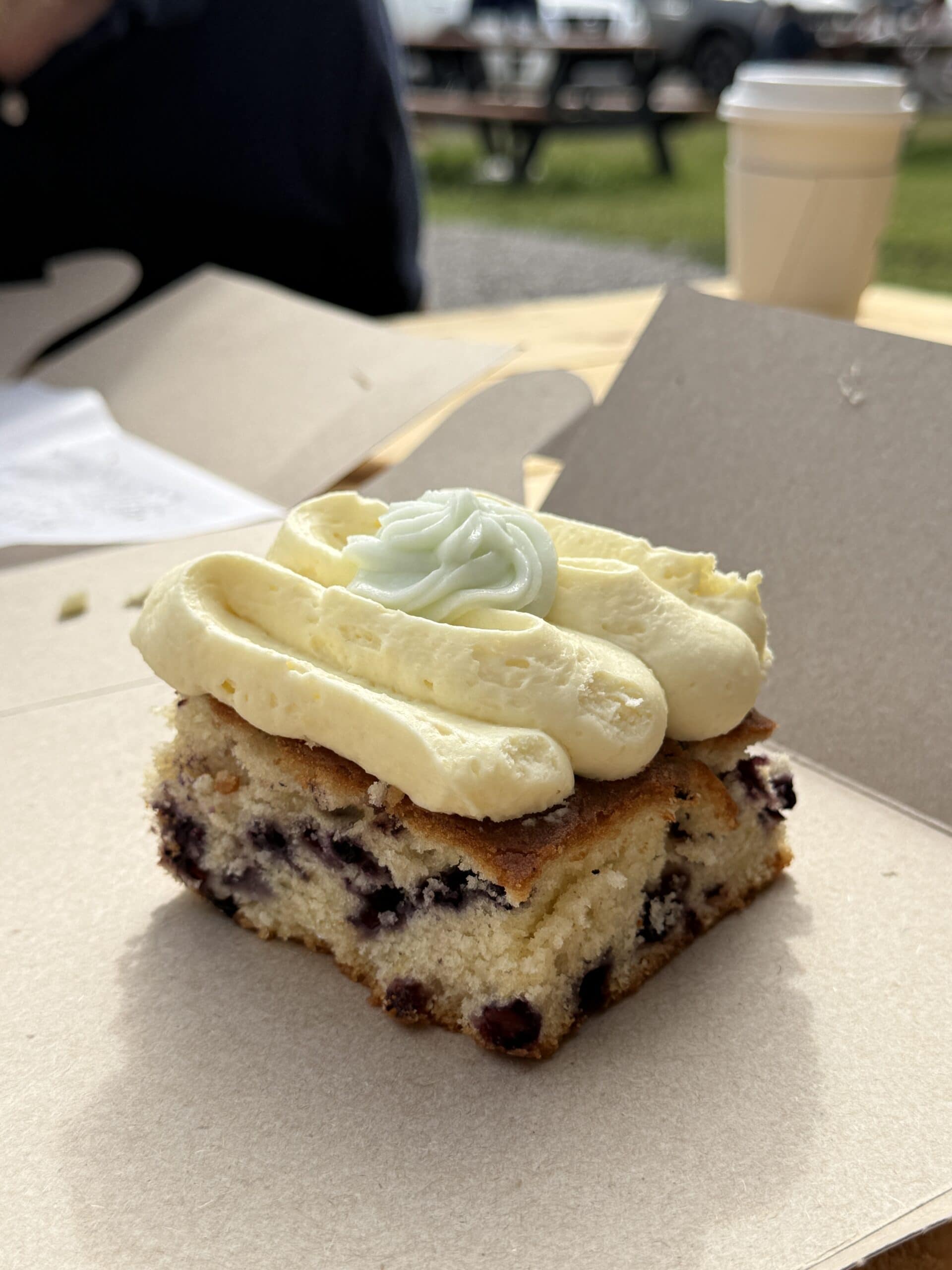 A square slice of blueberry cake with thick yellow frosting and a dollop of white cream sits on a cardboard tray. In the background, theres a paper coffee cup and outdoor seating.