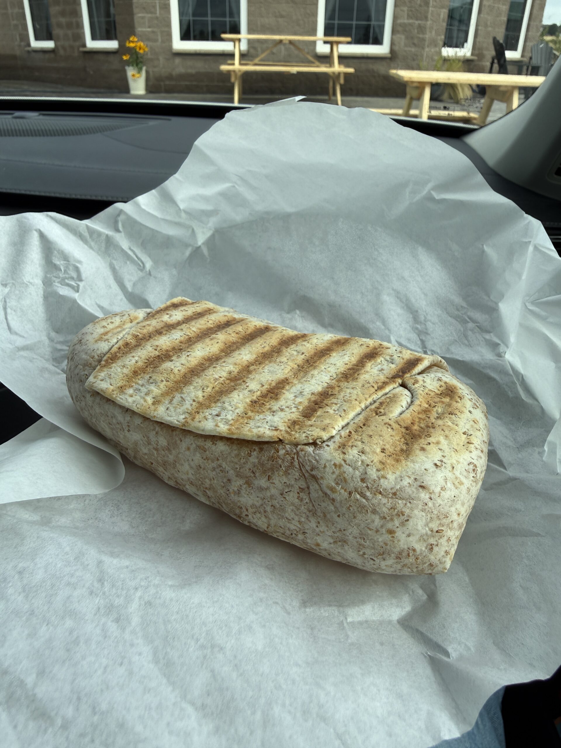 A large, grilled wrap sits on white parchment paper inside a car, with a picnic table and building visible outside the window in the background.