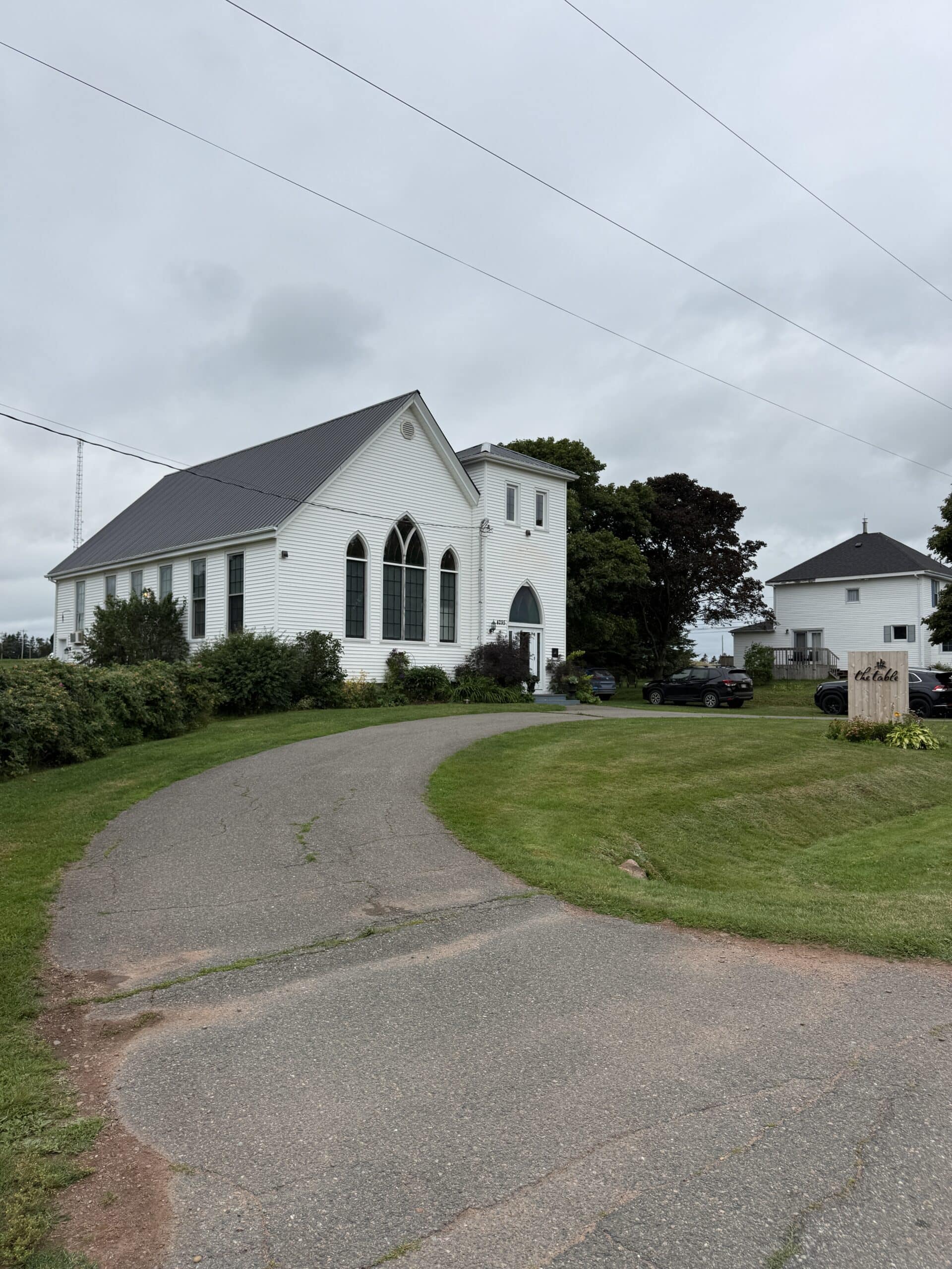 A white church with tall arched windows sits beside a curved driveway on a cloudy day. Green bushes, grass, and a few parked cars surround the building.