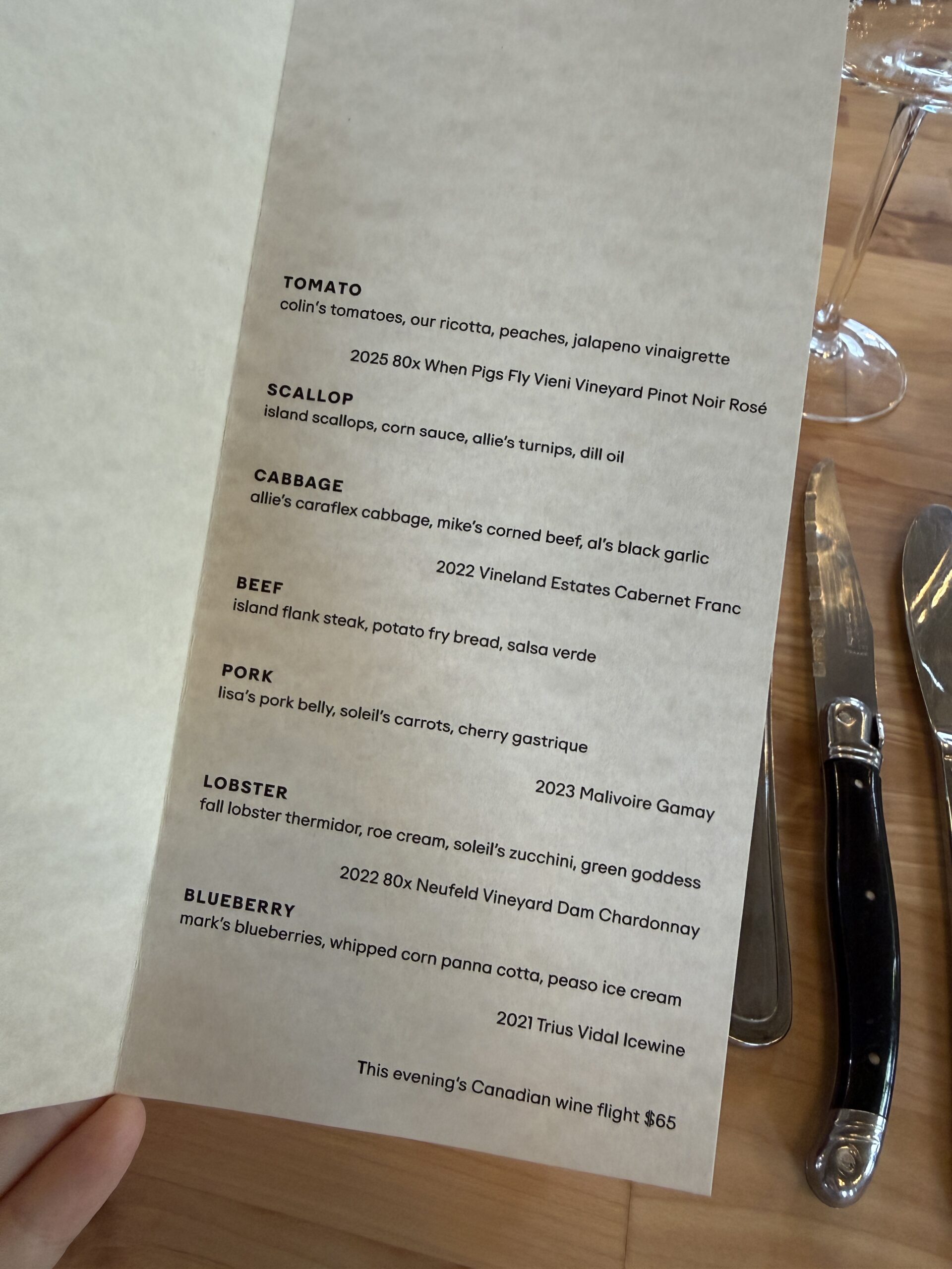 A dinner menu features dishes with tomato, scallop, cabbage, beef, pork, lobster, and blueberry, each paired with a specific wine. A fork, knife, and wine glass are set on a wooden table next to the menu.