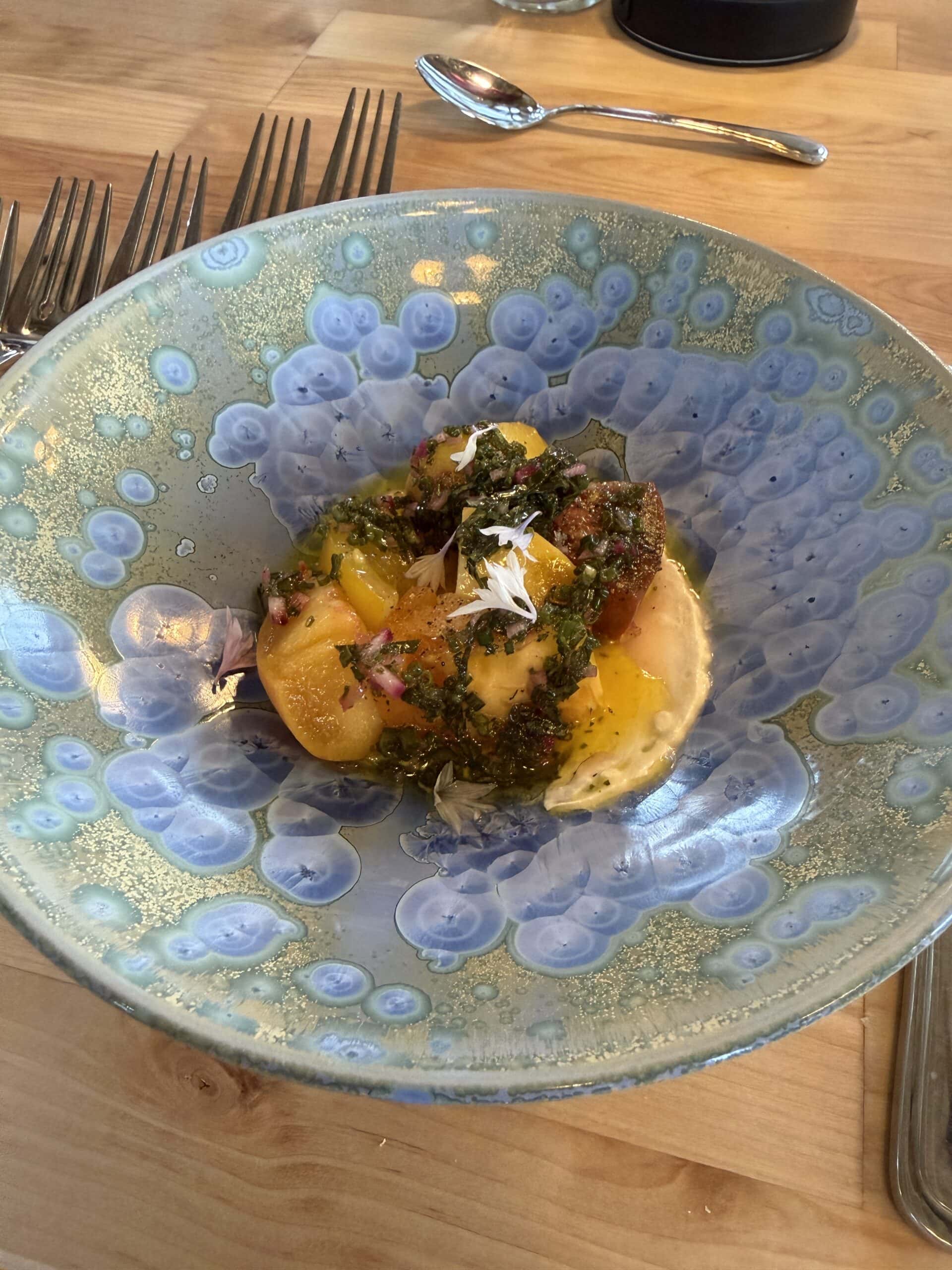 A blue ceramic bowl with a colorful salad, featuring sliced yellow and red tomatoes, herbs, and sauce, sits on a wooden table next to silverware.