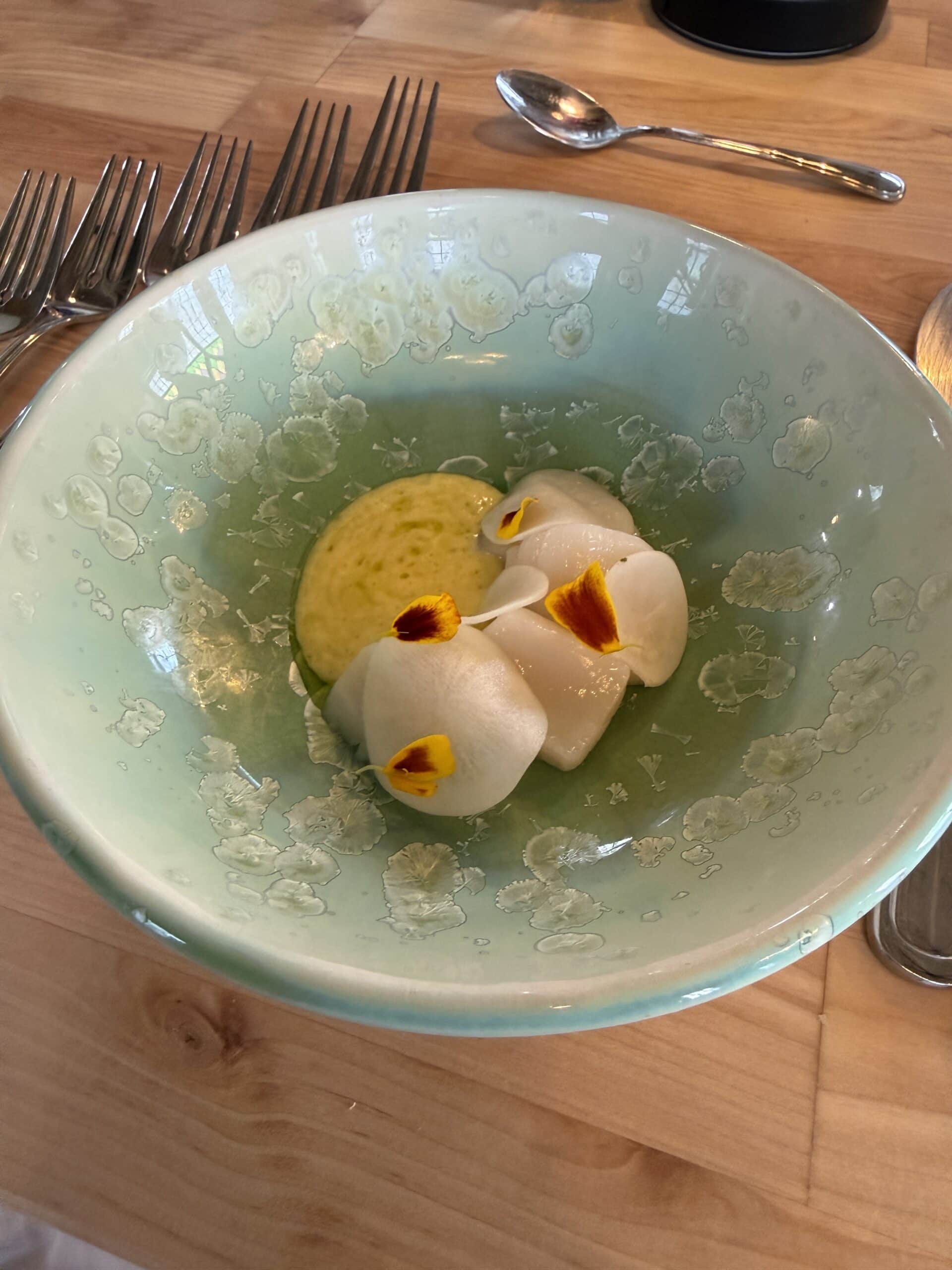A sea-green bowl with three white dumplings garnished with small yellow flowers, served with a green sauce. The bowl sits on a wooden table with silver forks and a spoon nearby.
