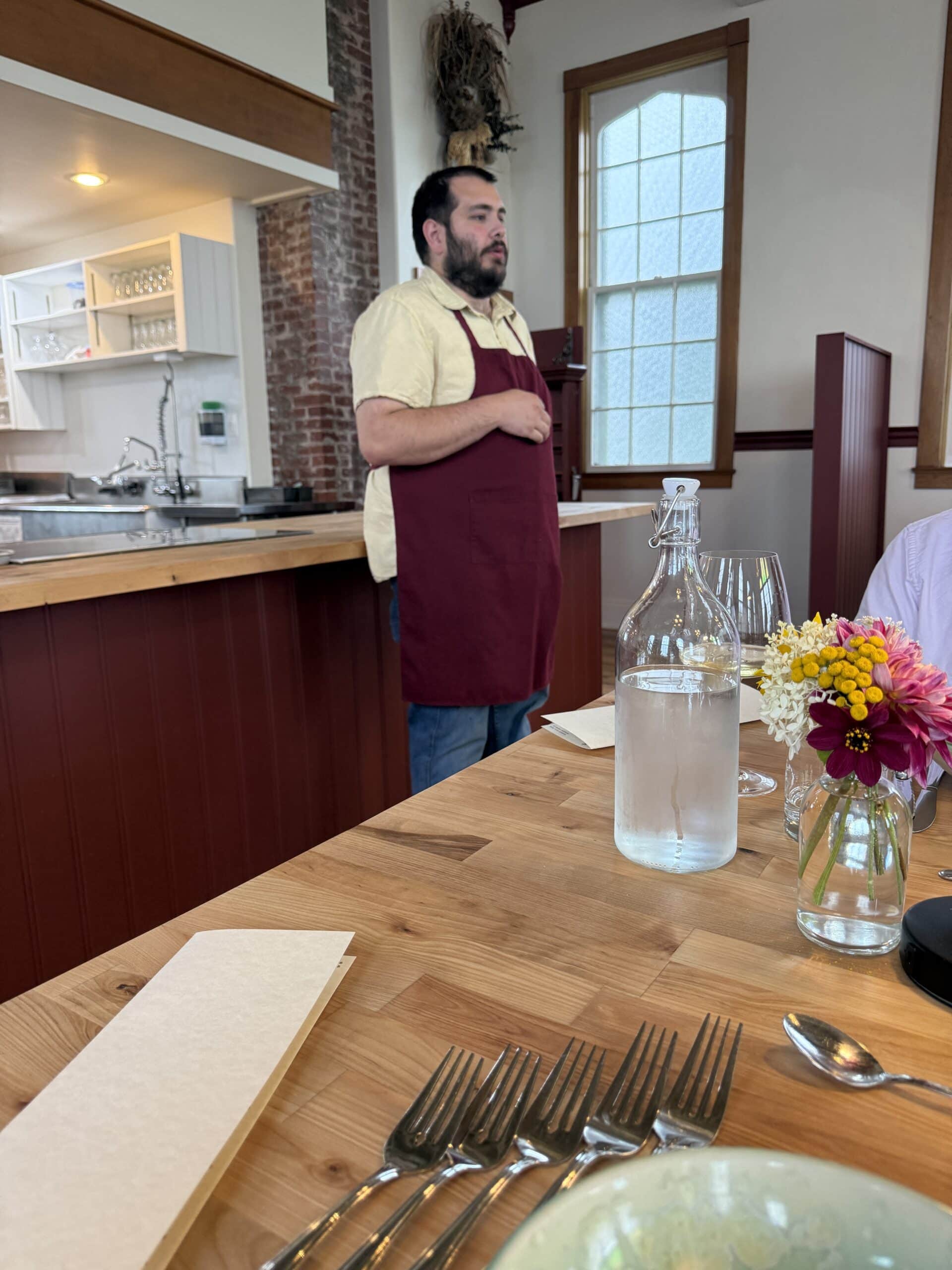 A man in a red apron stands by a wooden table set with silverware, a water bottle, wine glasses, napkins, and a small vase of flowers in a bright, rustic restaurant.