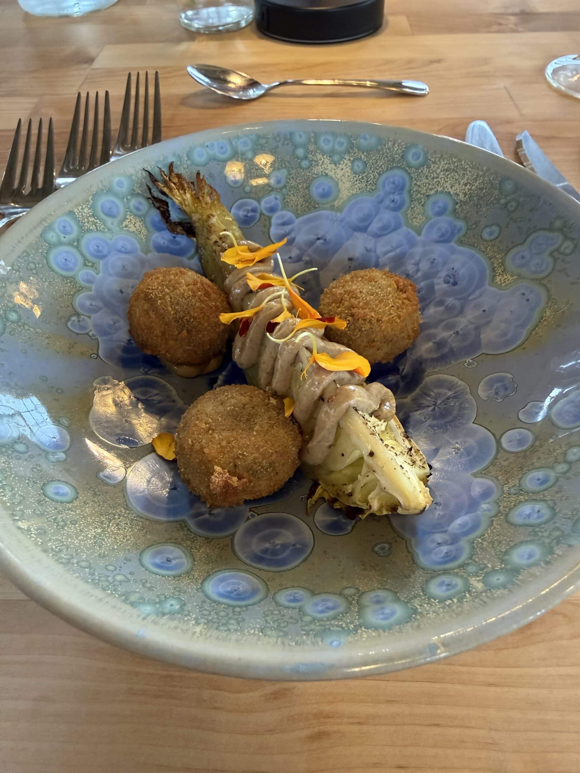 A ceramic plate with three breaded, fried balls next to grilled greens topped with a creamy sauce and yellow flower petals, on a wooden table with silverware in the background.
