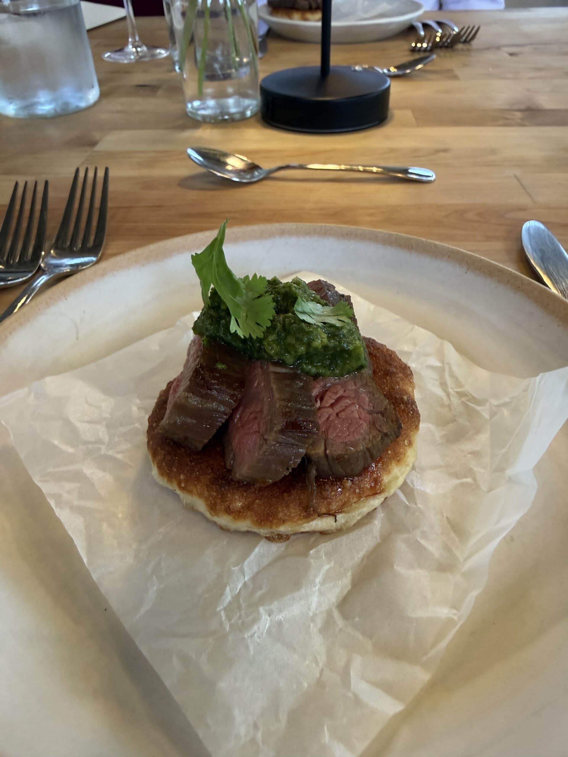 Slices of medium-rare steak topped with green sauce and herbs, served on a round base atop parchment paper on a plate. Forks and glasses are visible on the wooden table in the background.