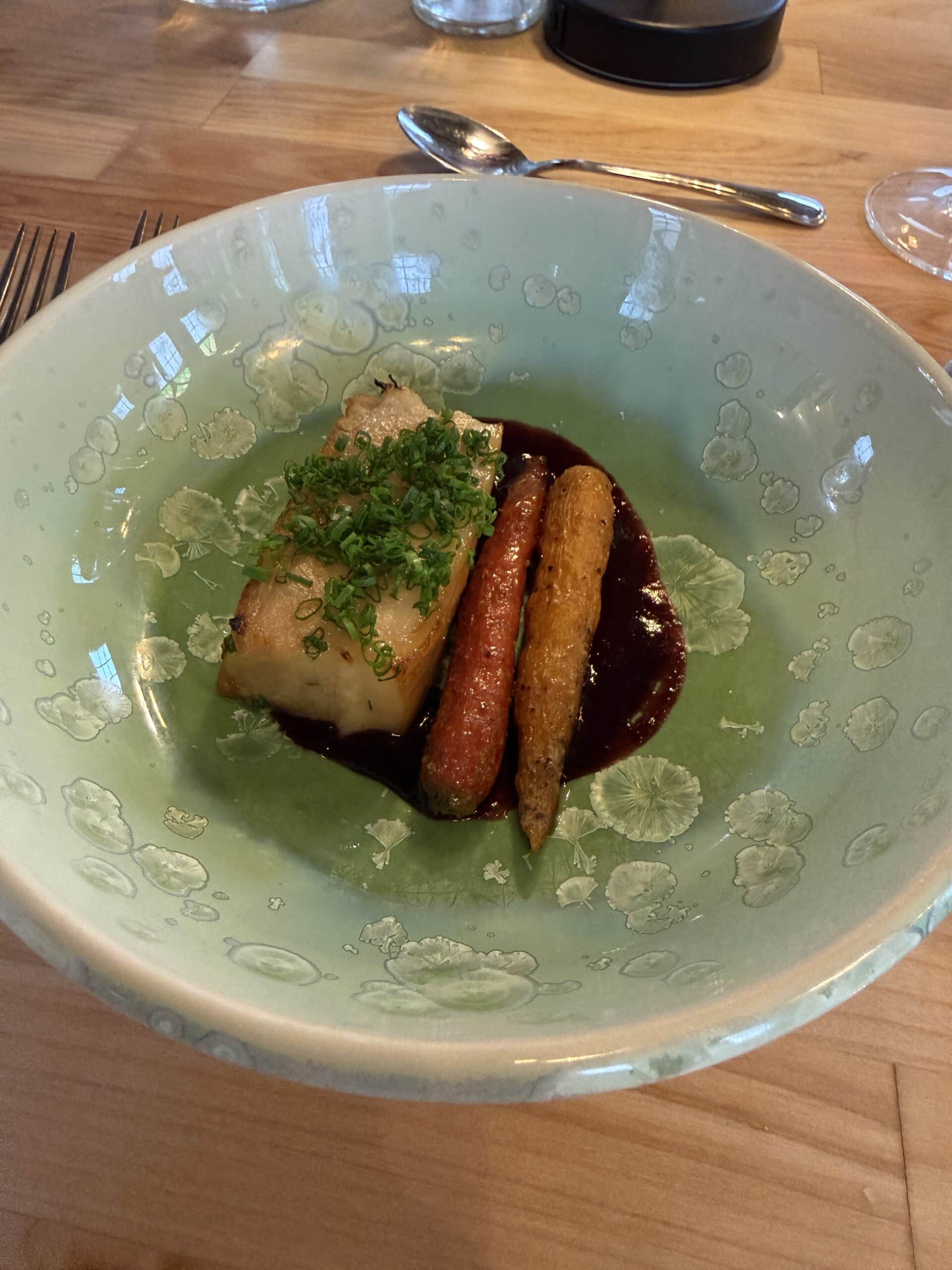 A green ceramic bowl with a piece of fish topped with herbs, served with two roasted carrots and a dark sauce, sits on a wooden table next to utensils and a glass.