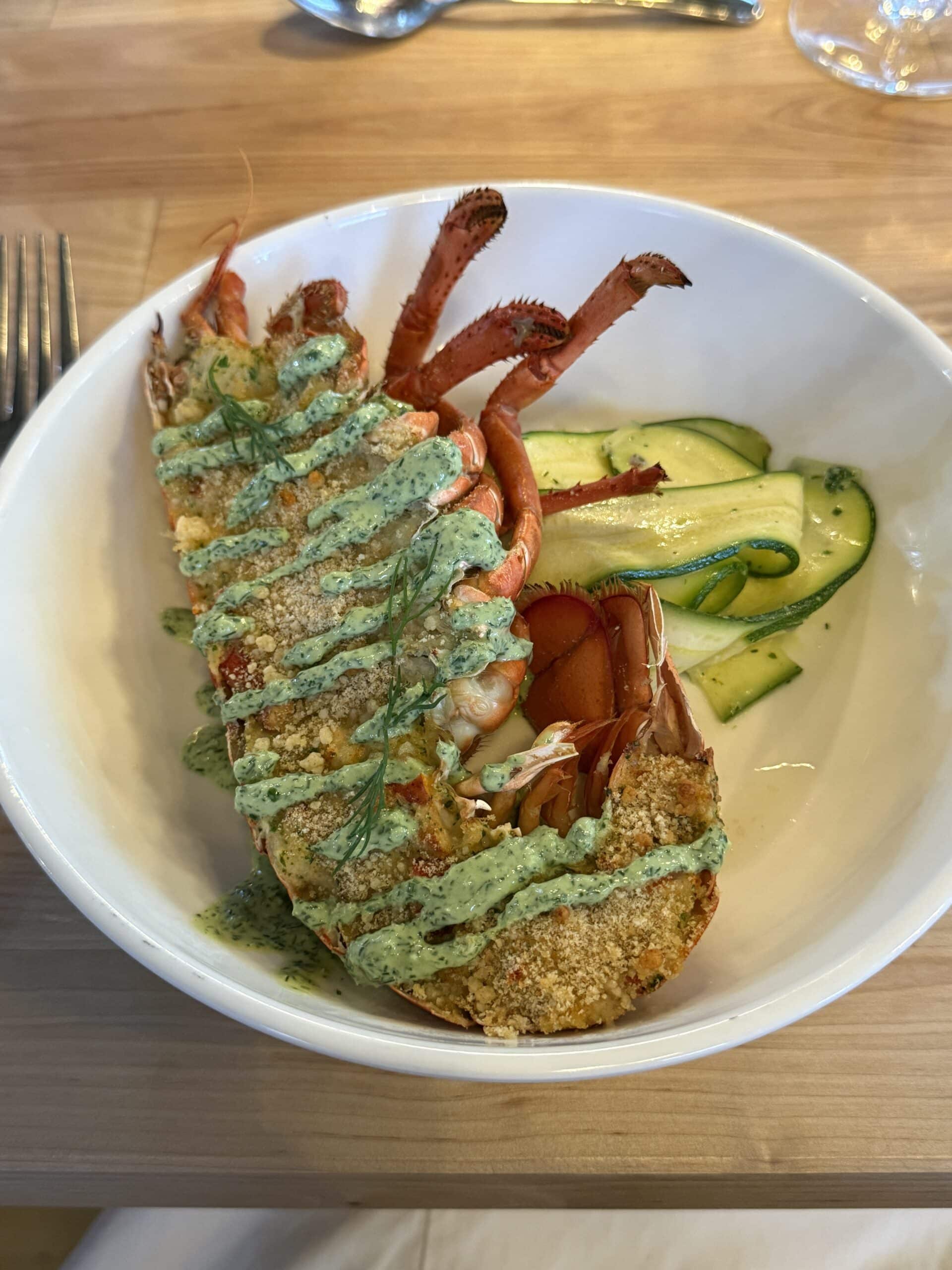 A baked lobster tail topped with green herb sauce and breadcrumbs, served in a white bowl with a side of ribboned zucchini on a wooden table.