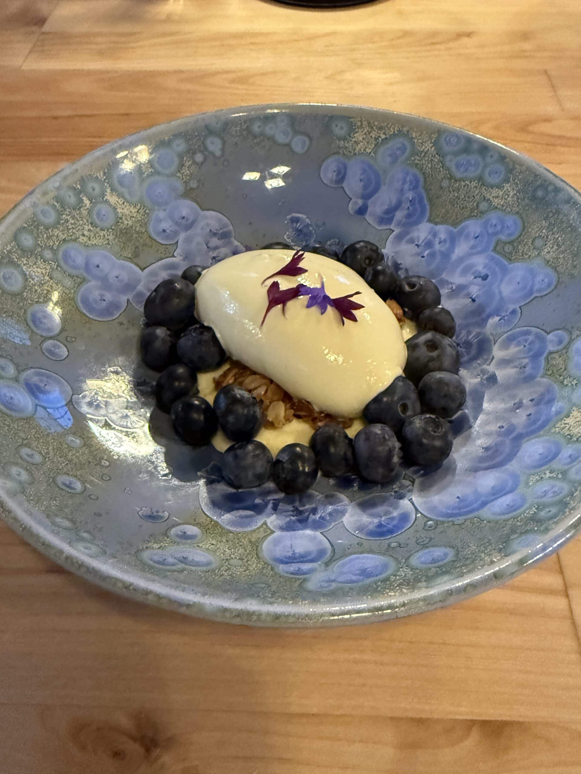 A blue ceramic plate with a ring of fresh blueberries, a dollop of cream or mousse in the center, garnished with purple flower petals, set on a wooden table.