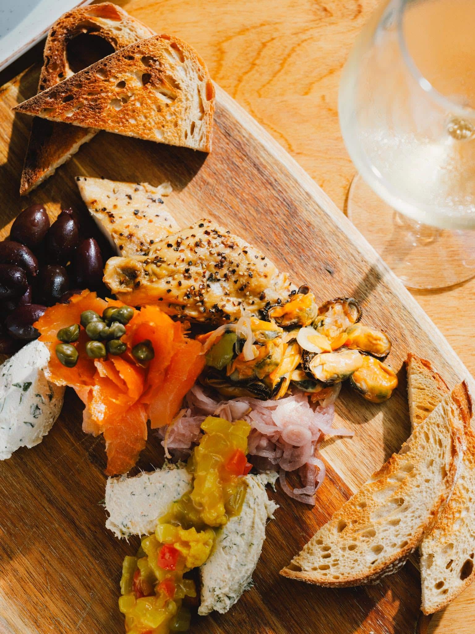 A wooden board with sliced toasted bread, olives, smoked salmon with capers, marinated seafood, herbed cheese, and a glass of white wine on the side.