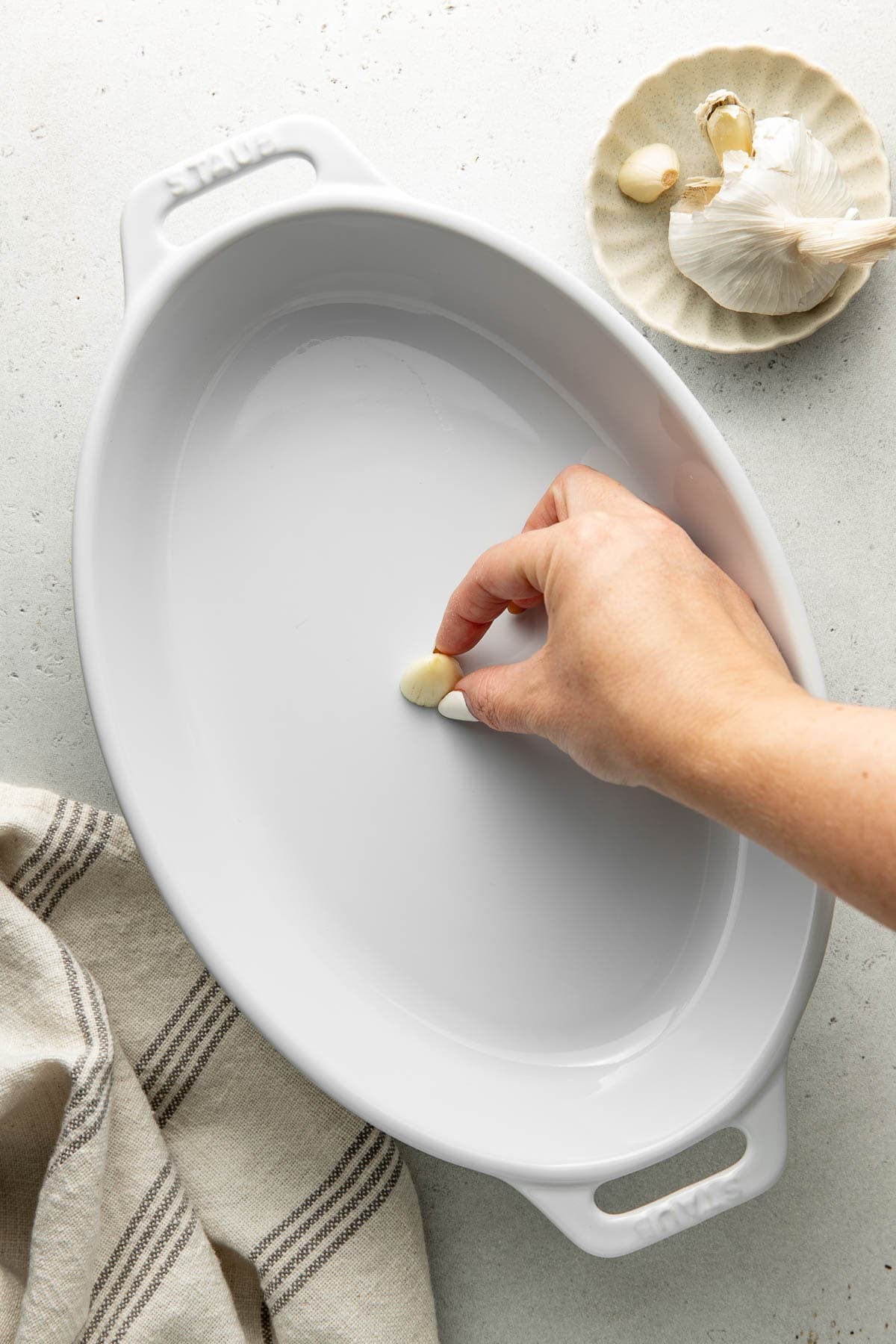 A hand rubs a halved garlic clove inside a white oval baking dish. A garlic bulb and cloves sit on a small plate nearby, with a striped kitchen towel partially visible.