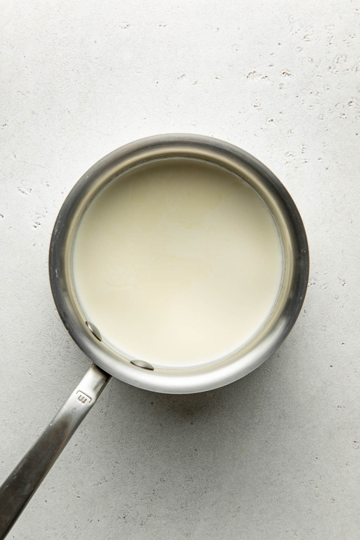 A stainless steel saucepan filled with milk, viewed from above, sits on a light gray textured surface.