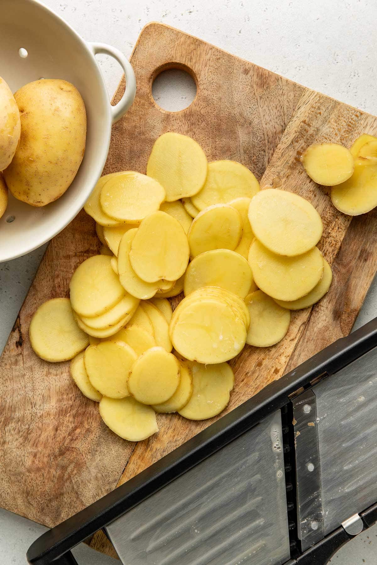 Thinly sliced potatoes are spread on a wooden cutting board, with a mandoline slicer nearby and a white colander holding whole potatoes beside them.