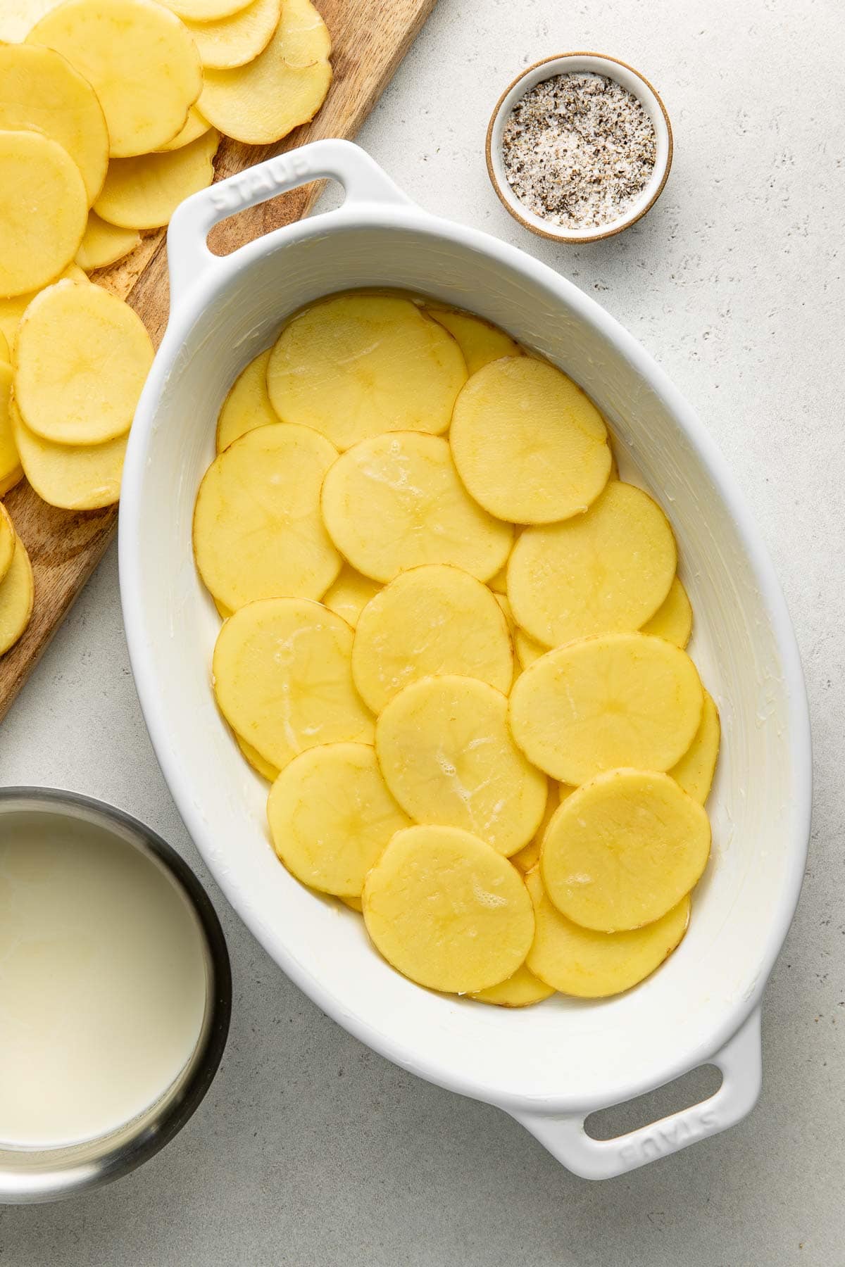 Sliced potatoes layered in a white oval baking dish, with a bowl of milk, a wooden board with more potato slices, and a small bowl of salt and pepper on a light surface.