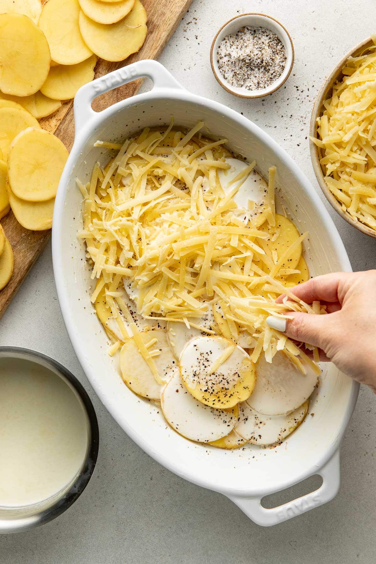 A hand sprinkles shredded cheese on sliced potatoes layered in a white baking dish with a creamy sauce, black pepper, and more cheese and sliced potatoes nearby.
