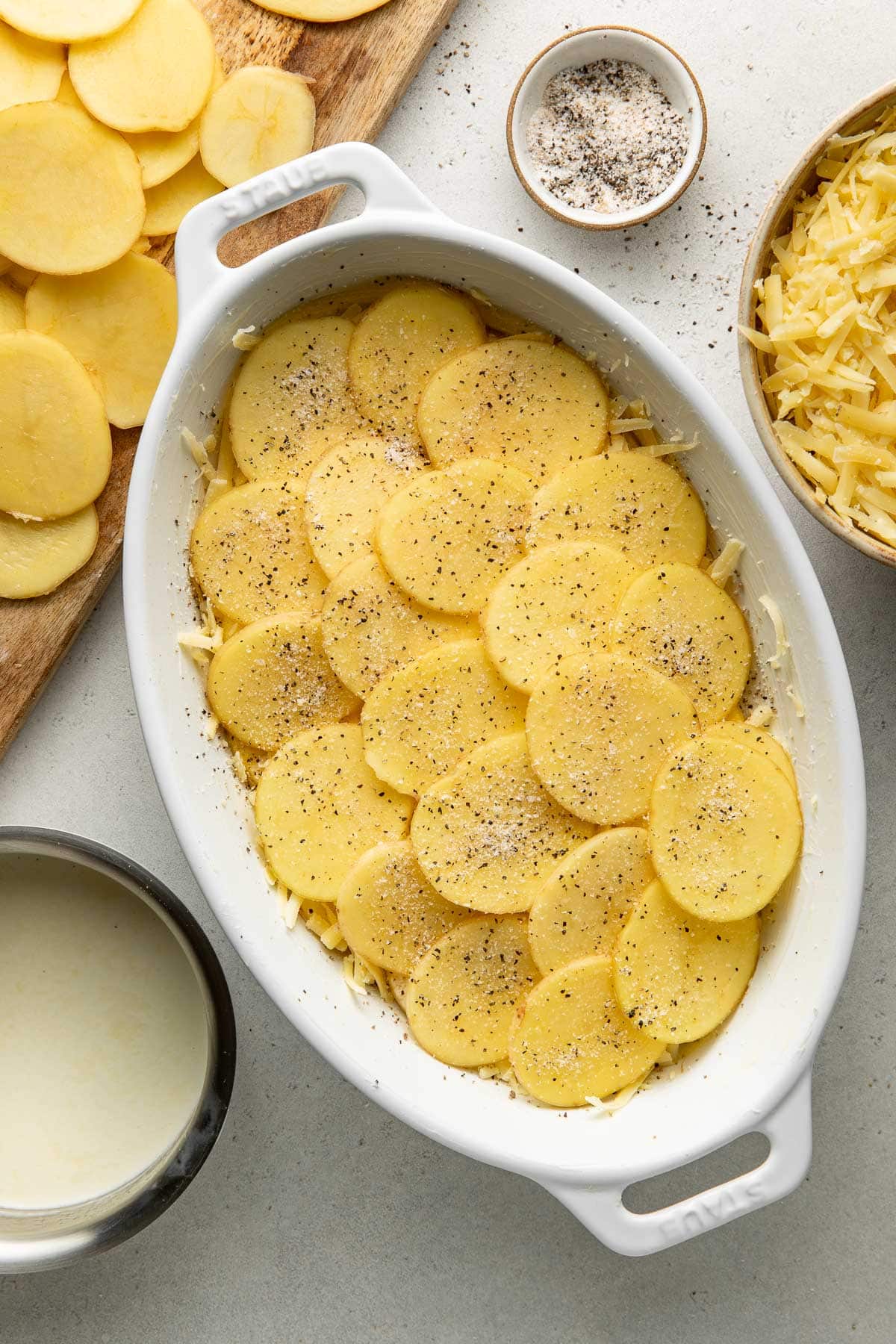 A white baking dish filled with sliced potatoes, sprinkled with black pepper. Surrounding the dish are a bowl of shredded cheese, a cup of cream, a cutting board with sliced potatoes, and a small bowl of pepper.