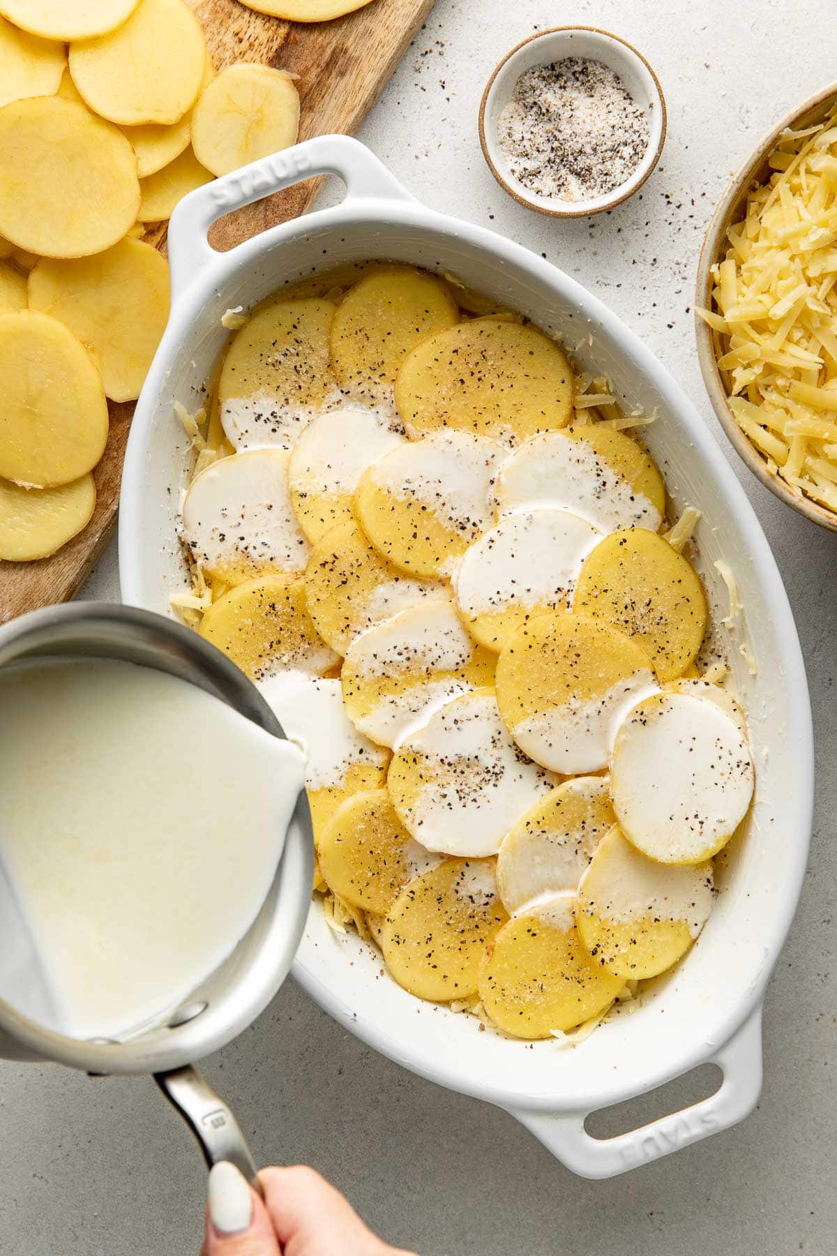 Sliced potatoes in a white baking dish being covered with cream from a saucepan, with grated cheese, seasoning, and more potato slices nearby.