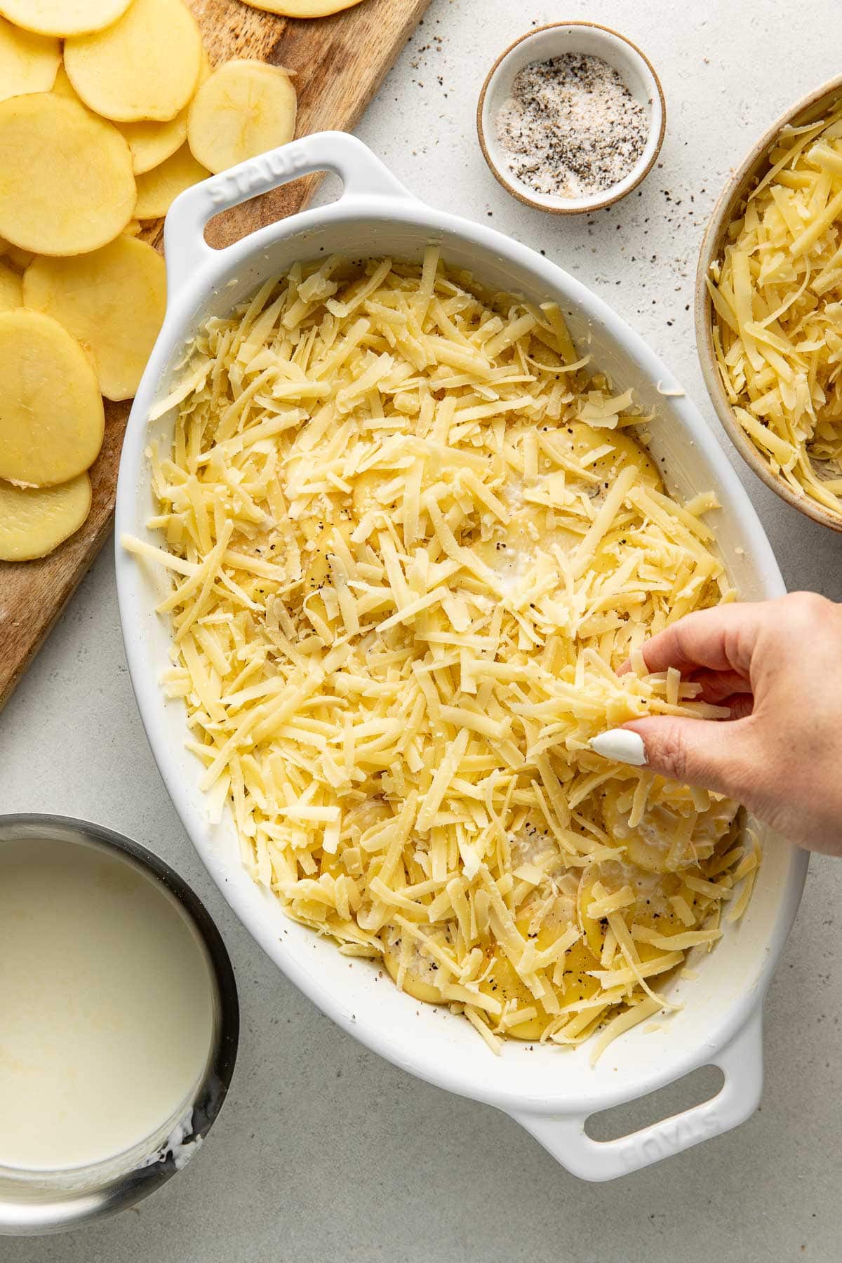 A hand sprinkles shredded cheese over sliced potatoes in a white baking dish, with a bowl of milk, a small bowl of seasoning, and sliced potatoes on a cutting board nearby.