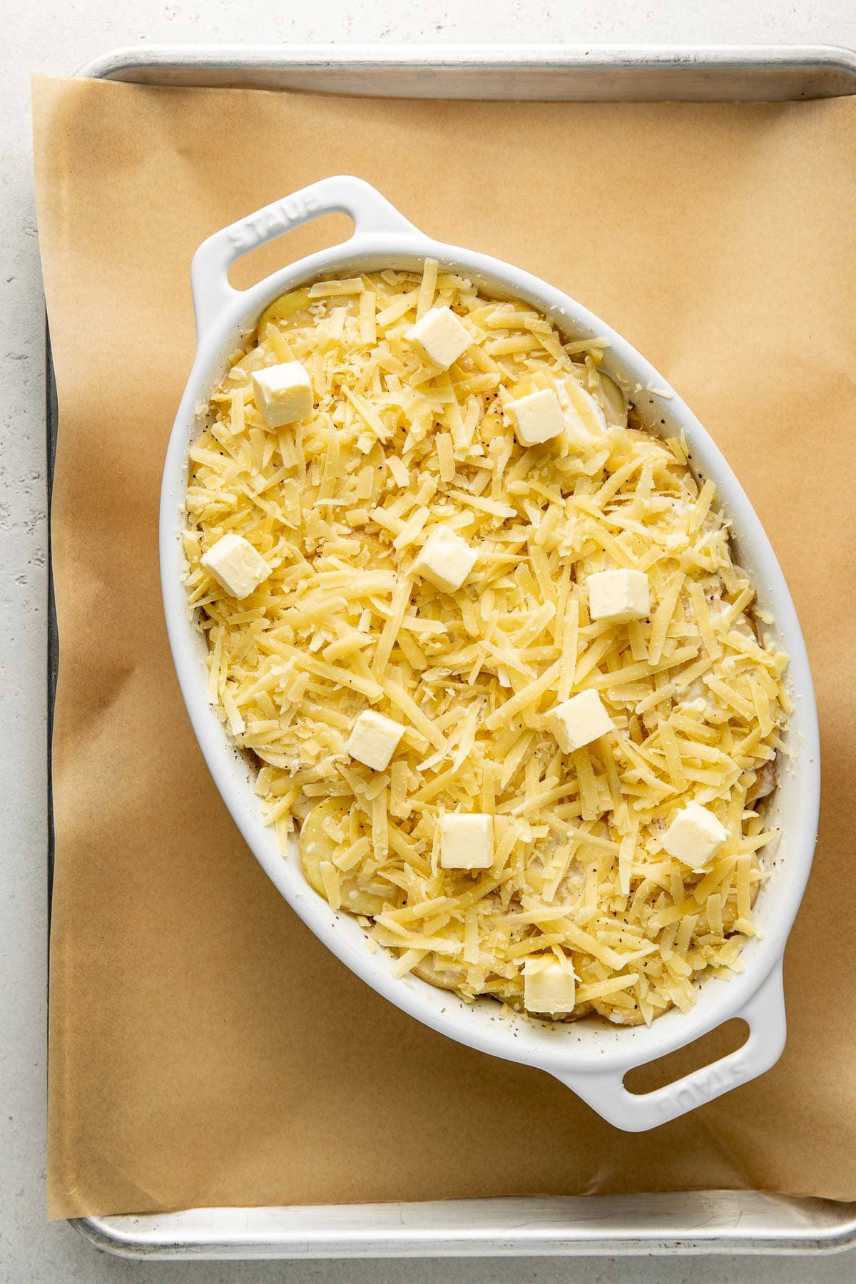 Oval white baking dish filled with shredded cheese and topped with small cubes of butter, placed on a baking sheet lined with parchment paper.