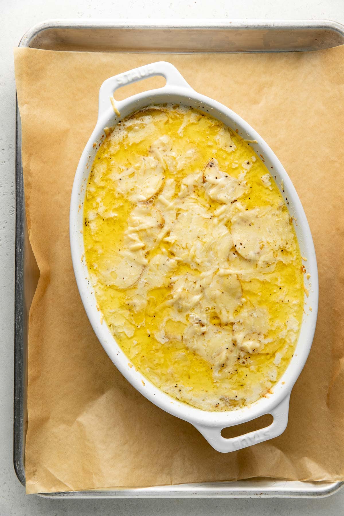 An oval white baking dish filled with cheesy, golden-brown scalloped potatoes sits on parchment paper on a metal baking sheet.
