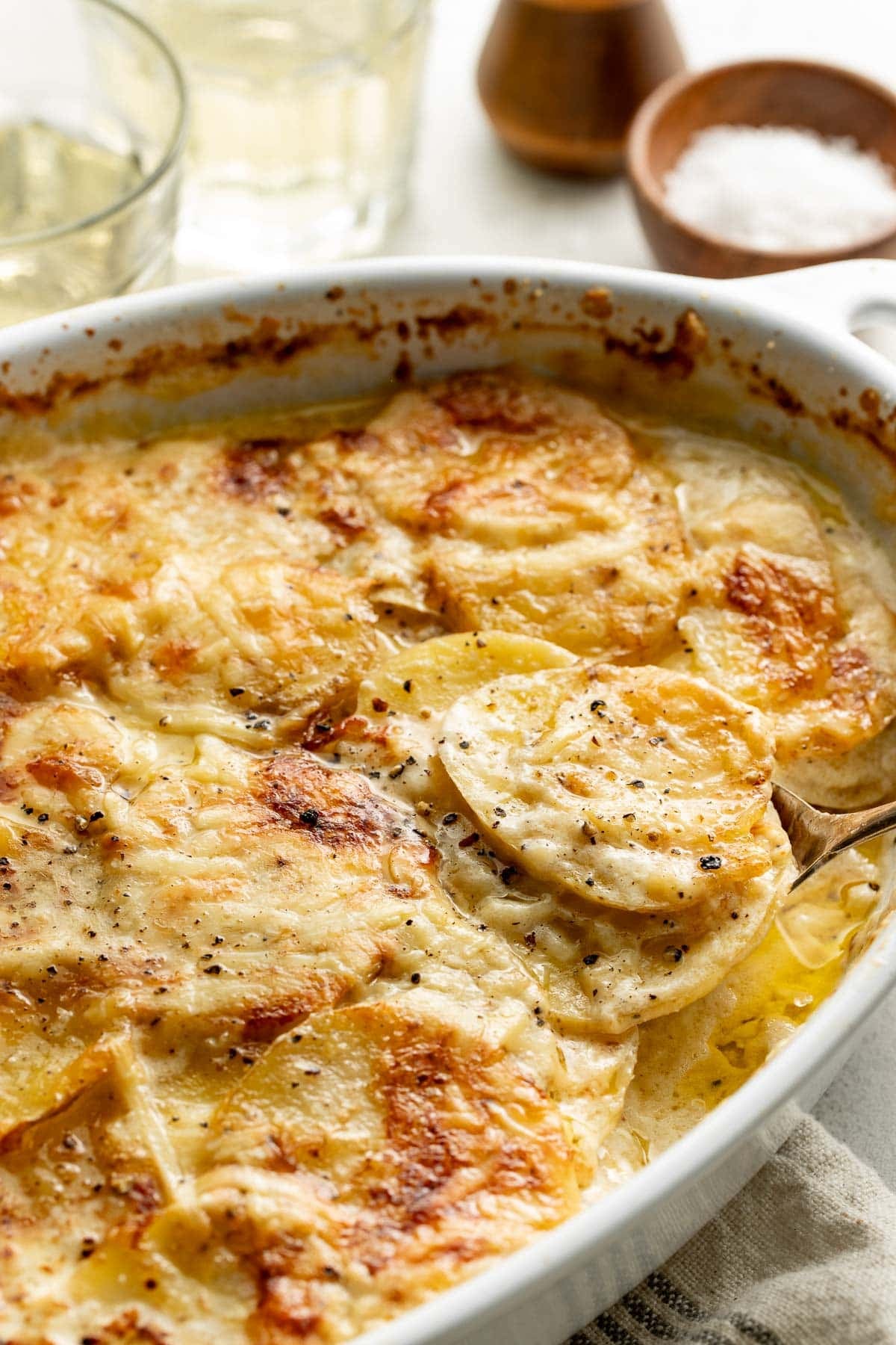 Close-up of a baked dish of scalloped potatoes with melted cheese and golden-brown crust, served in a white ceramic baking dish, with a spoon lifting a portion. Glasses and a salt bowl are in the background.
