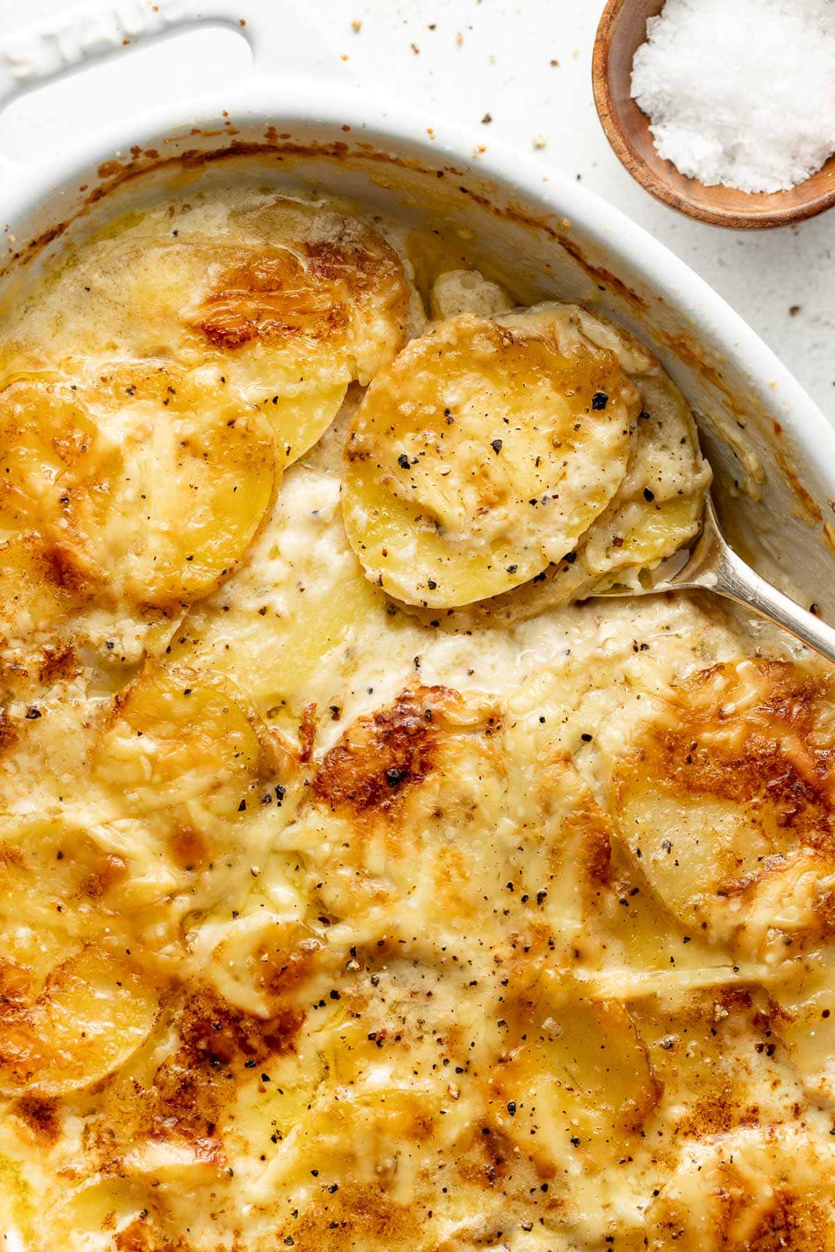 Golden-brown scalloped potatoes in a creamy sauce, partially served with a spoon, in a white baking dish. A small wooden bowl of flaky sea salt is visible in the top right corner.
