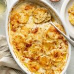 An oval baking dish of golden, cheesy scalloped potatoes, with a spoon scooping some out. A plate with a serving, salt, pepper, and striped napkin are nearby on a light surface.